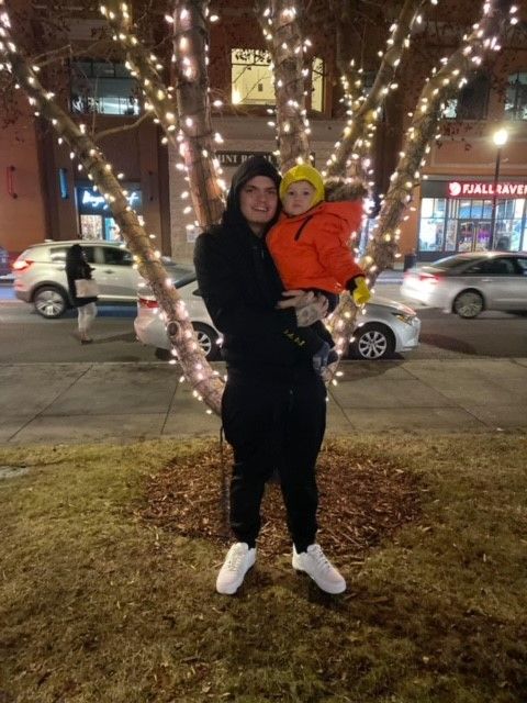 A man is holding a child in front of a tree with lights on it.