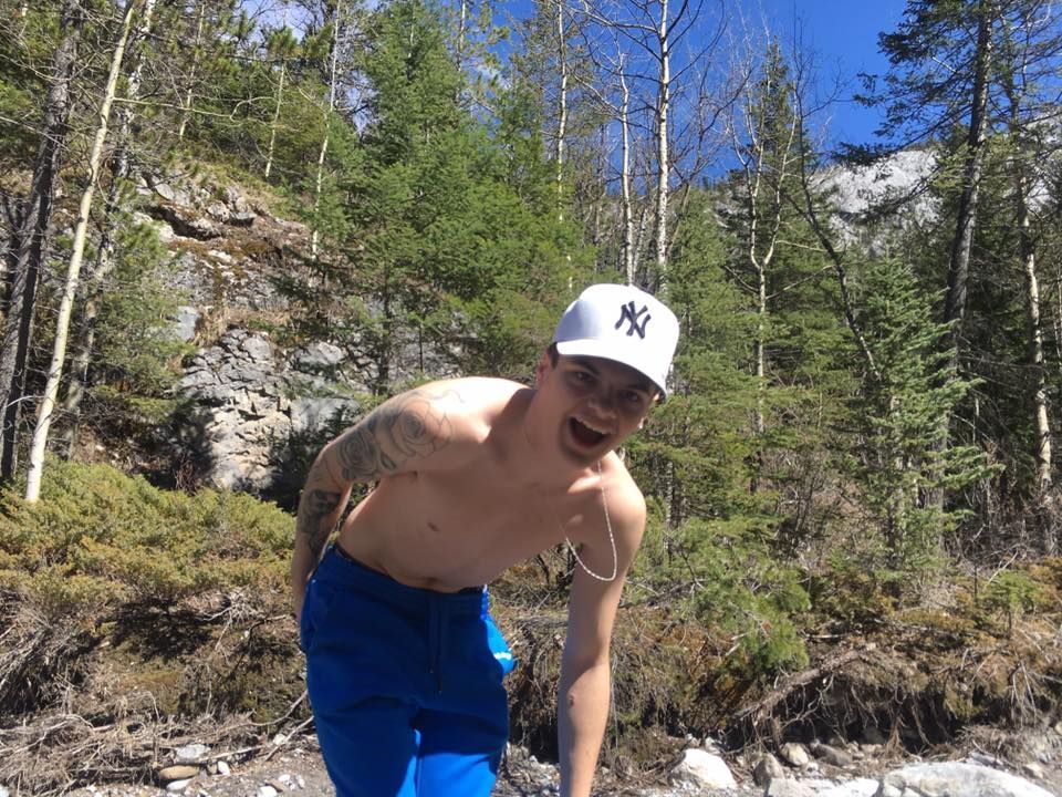 A shirtless man wearing a ny hat and blue shorts is standing in the woods.