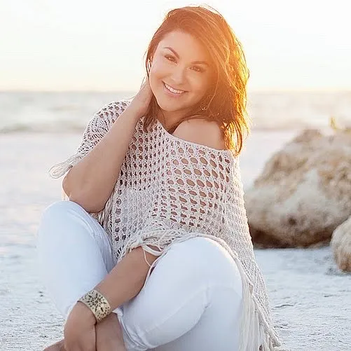 A woman in a crocheted top and white pants is kneeling on the beach.