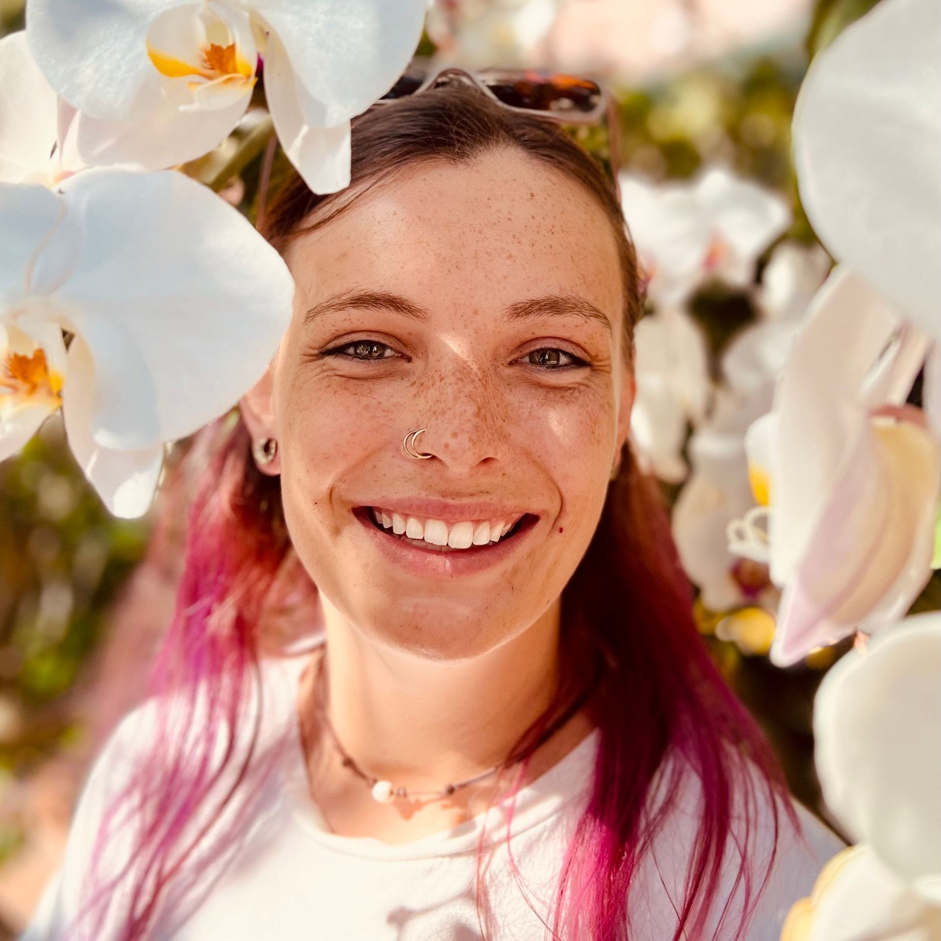 A woman with pink hair is smiling in front of flowers.