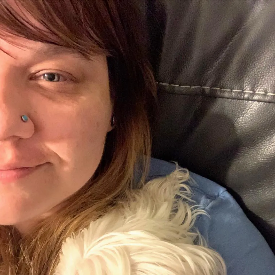 A woman with a nose ring is laying on a couch with a white dog.