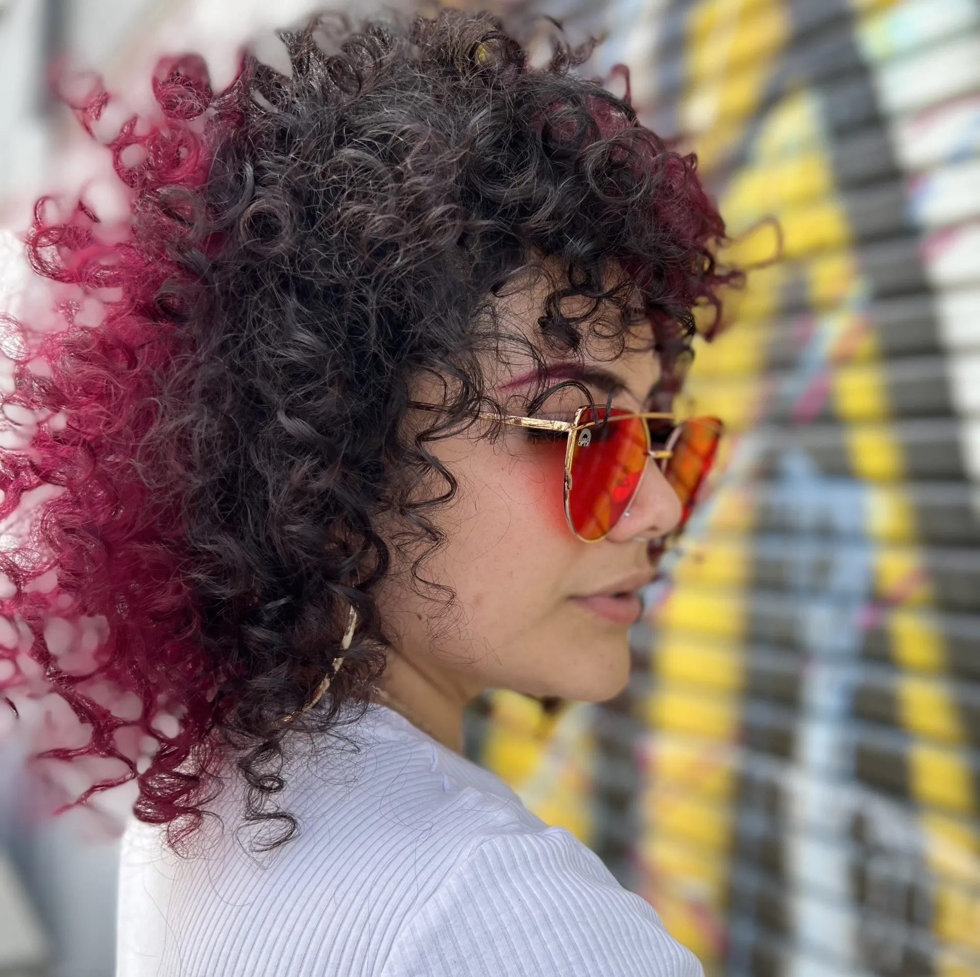 A woman with curly hair and sunglasses is standing in front of a graffiti wall.