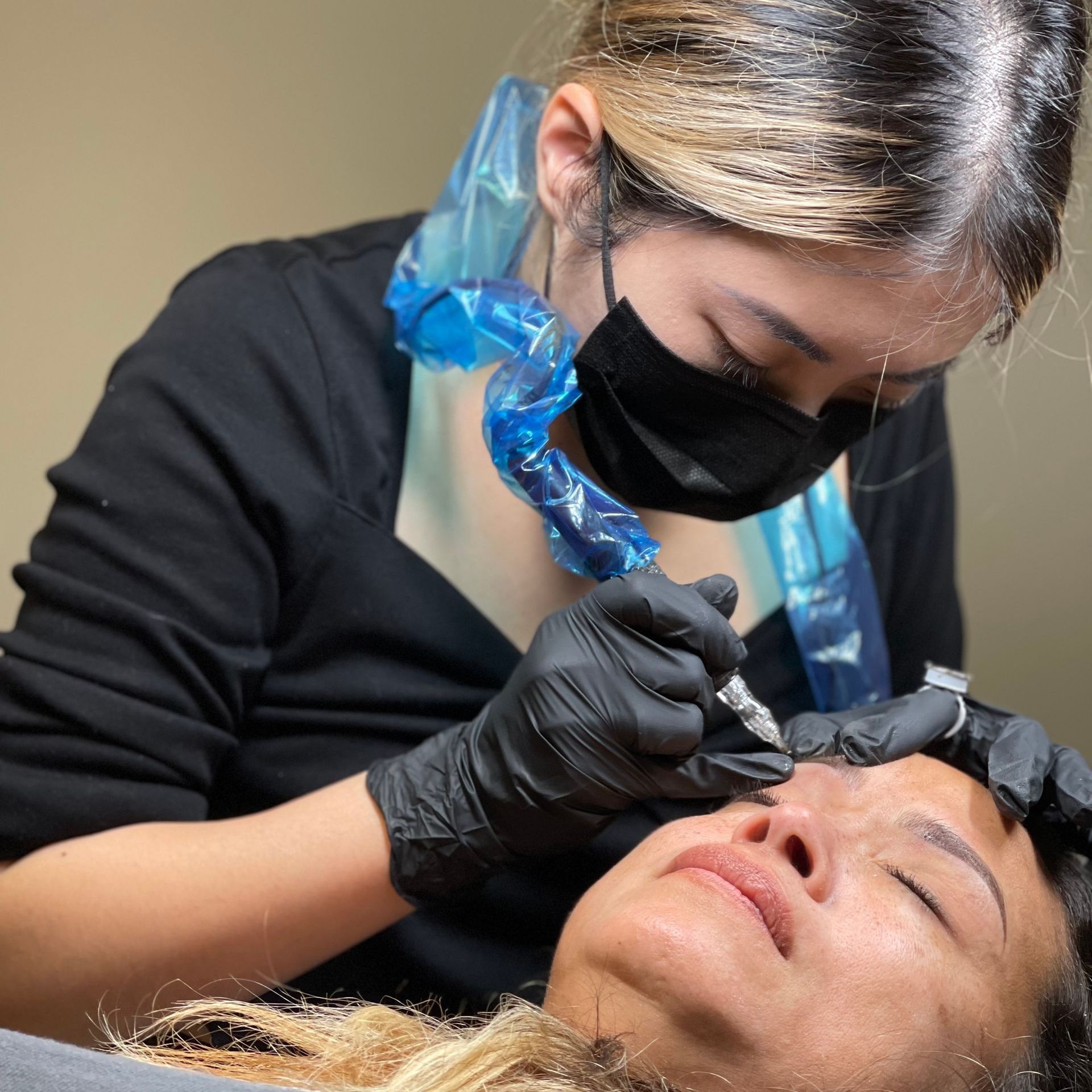 A woman wearing a mask and black gloves is tattooing a woman 's eyebrows