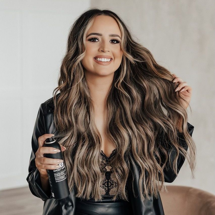 A woman with long hair is holding a bottle of hair spray and smiling.