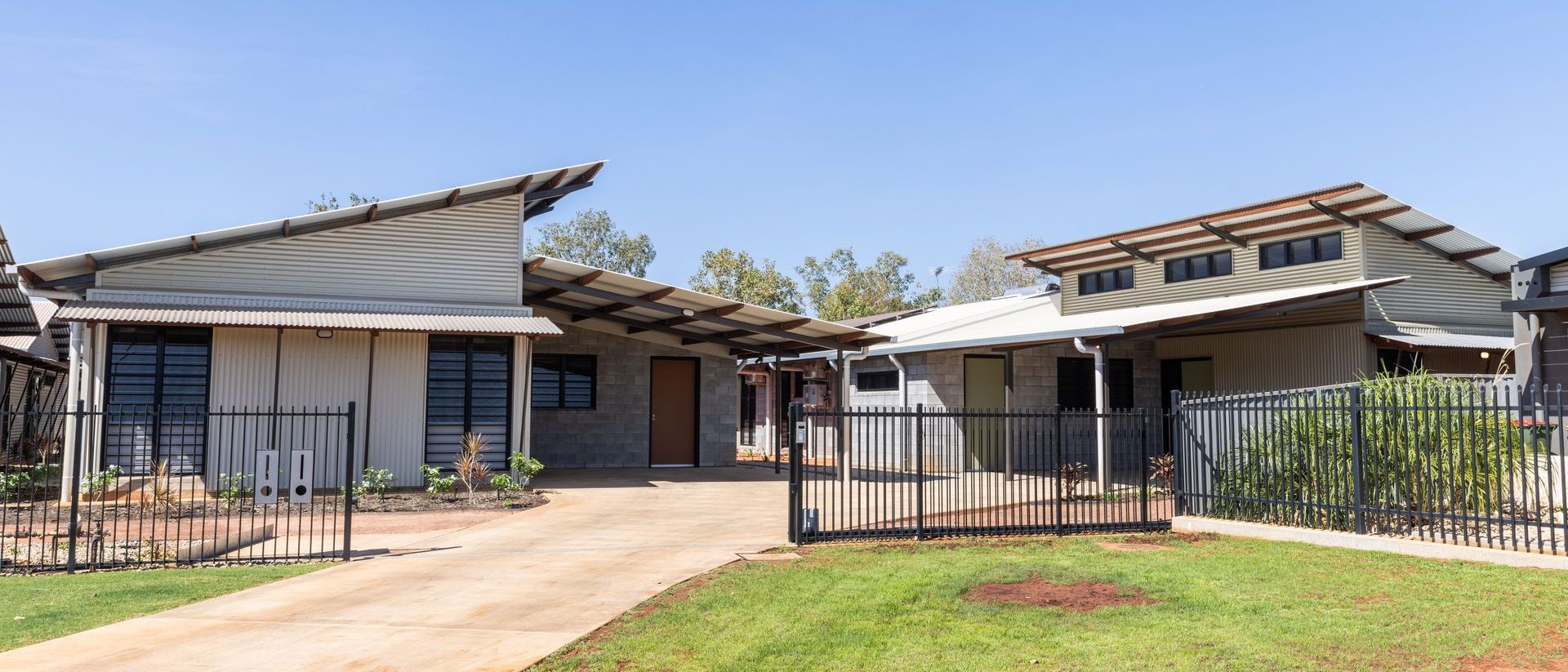 Houses with angled tin roofs, concrete drive, and green lawn. Black iron fence and gate.