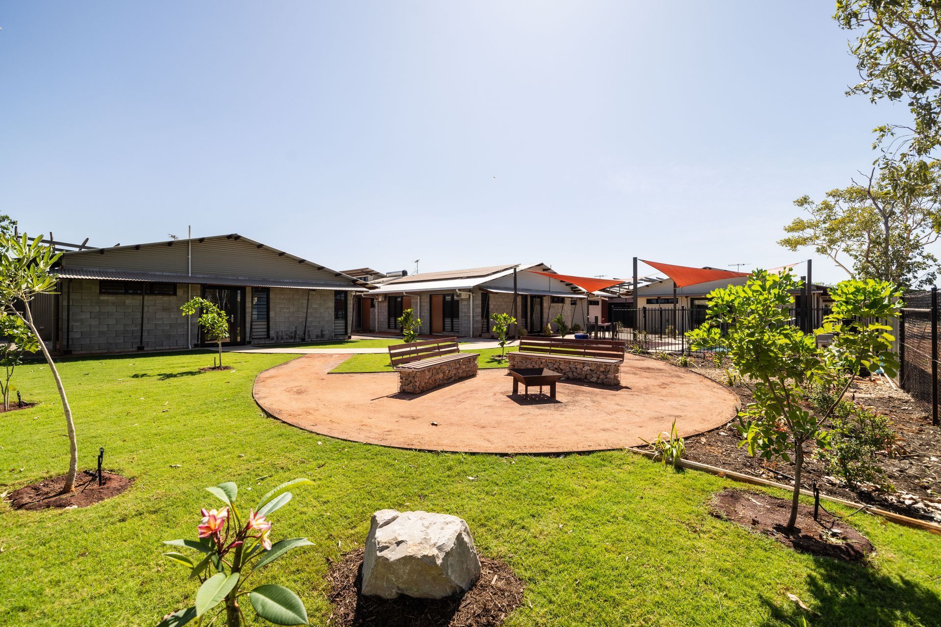 Grassy backyard with buildings, gravel seating area, benches, and young trees under a blue sky.