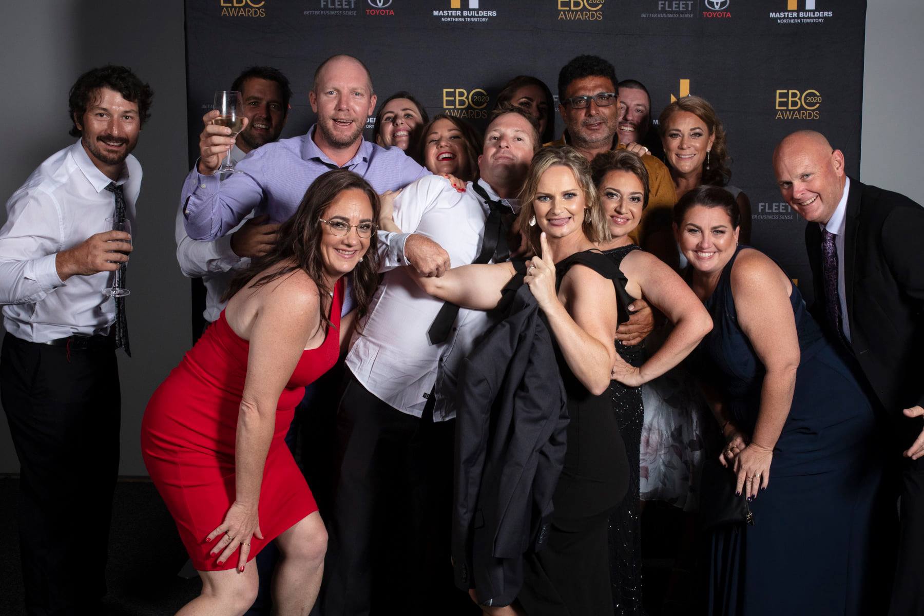 Group of people at an event, smiling and posing in front of a black backdrop. Some are holding drinks.