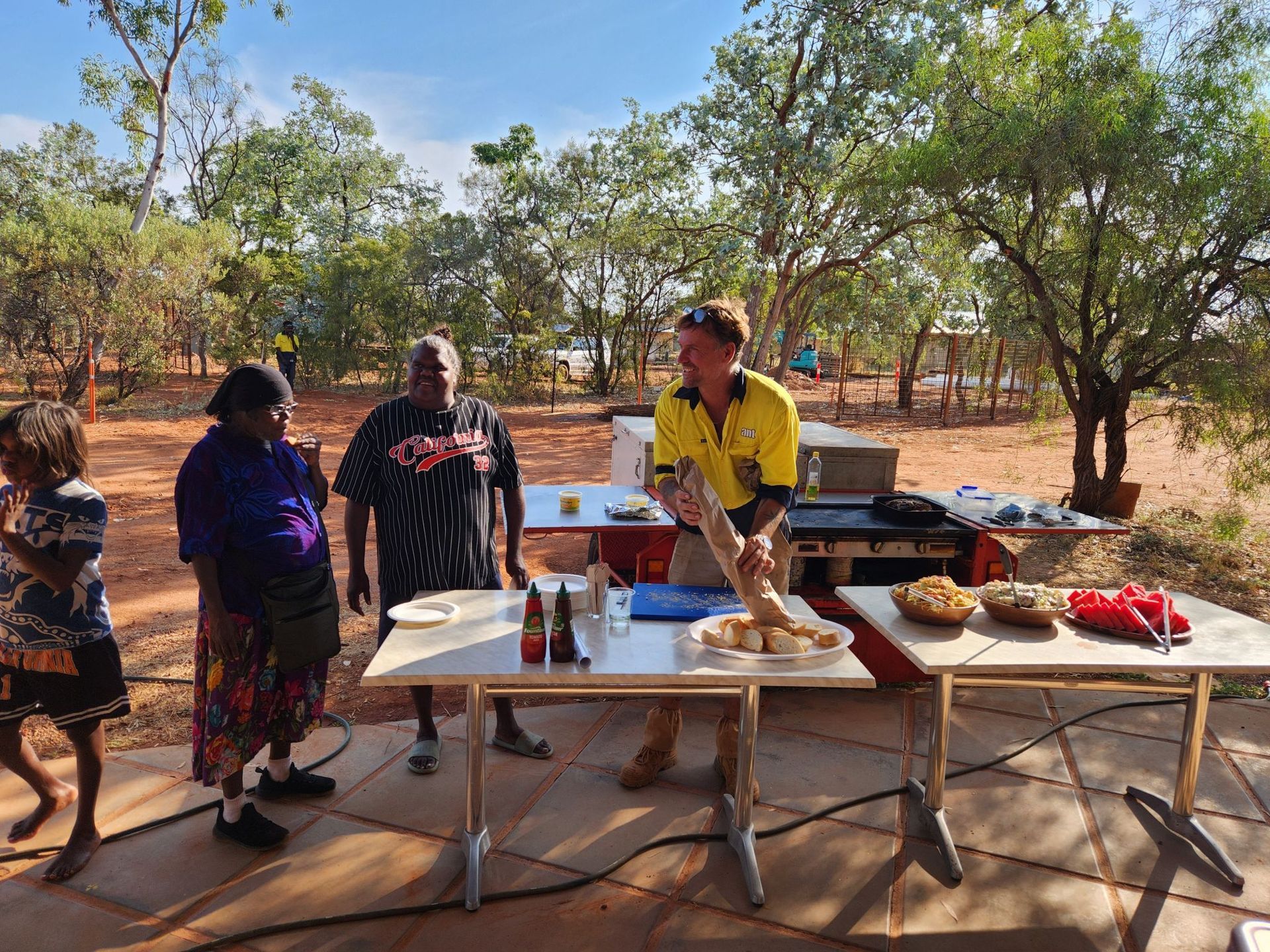 Members of community and M+J team member smiling at a barbeque with the red dirt landscape behind them
