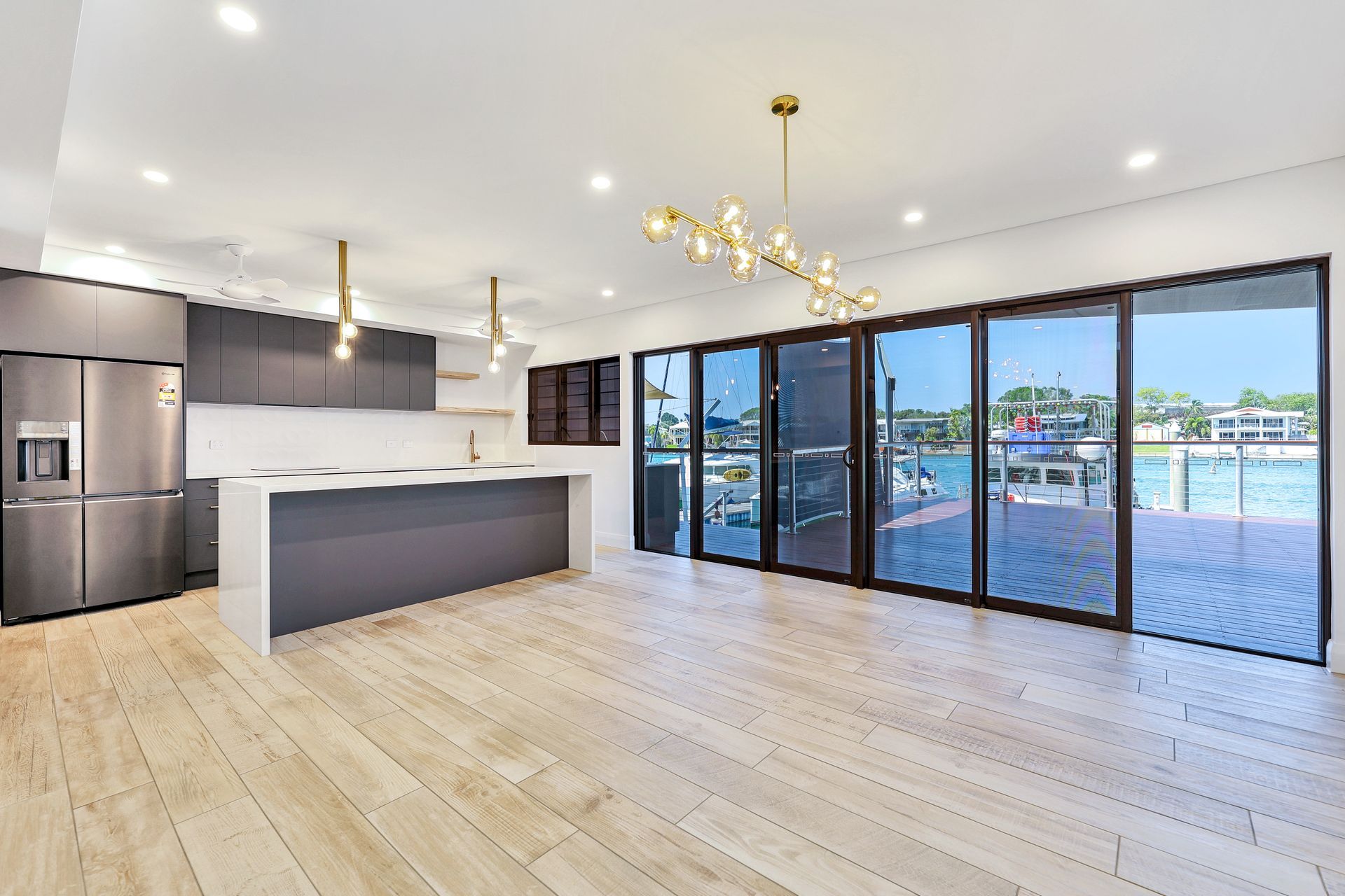 Open-plan modern kitchen with gray cabinets, white countertops, and sliding glass doors leading to a waterfront view.