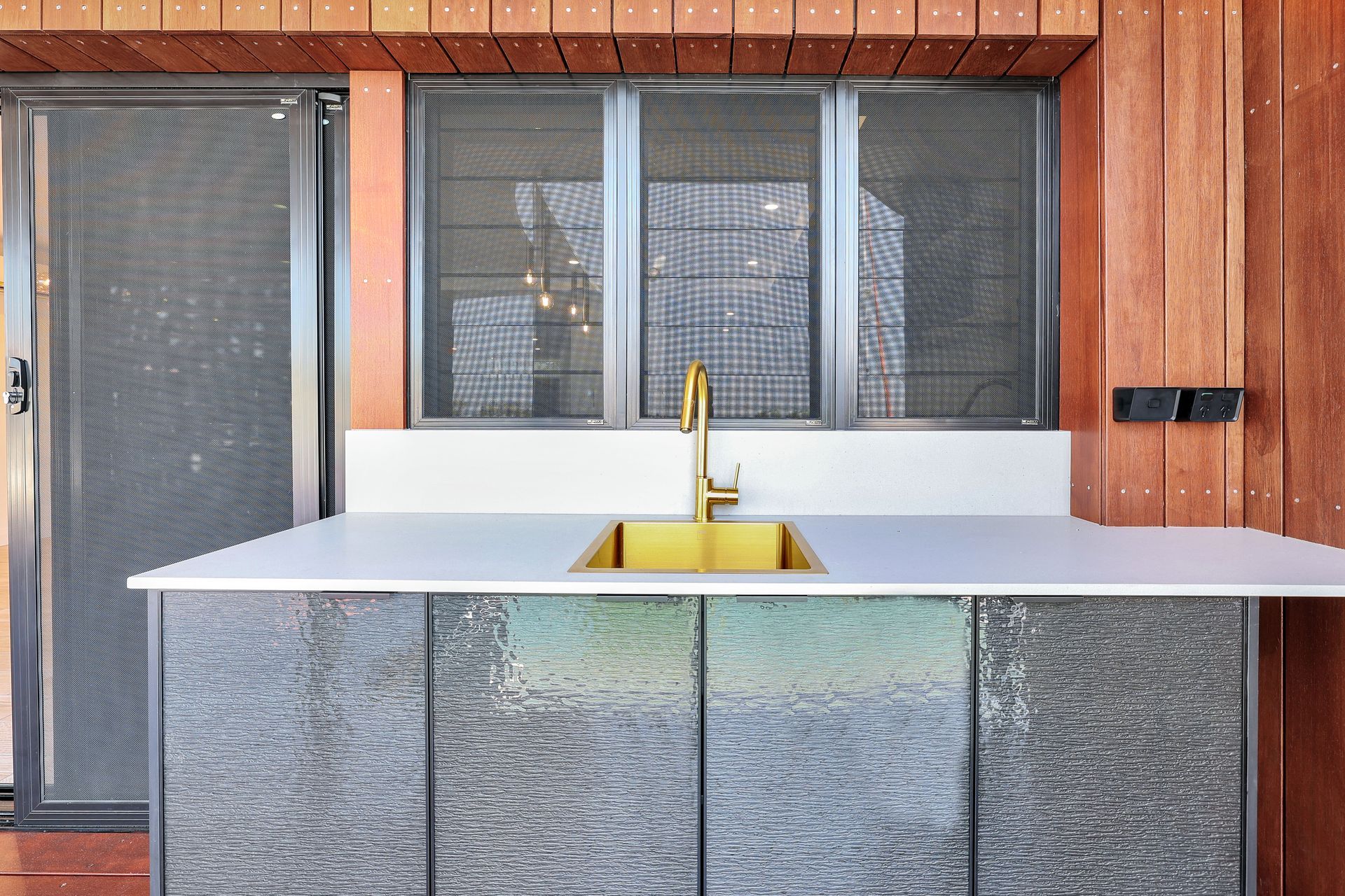 Outdoor kitchen with gold faucet, white counter, gray cabinets, wooden walls.