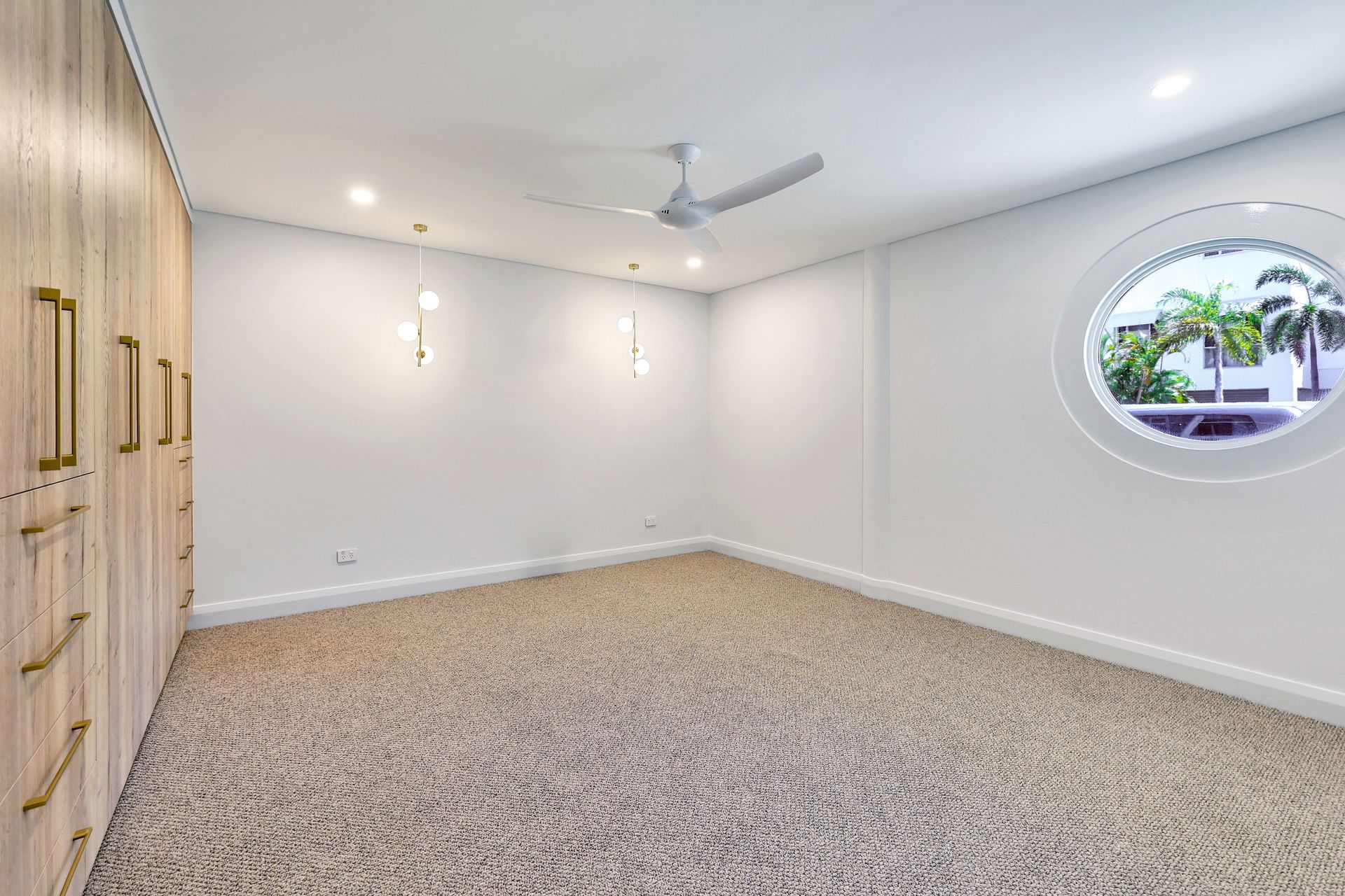 Empty bedroom with beige carpet, white walls, built-in closet, round window, and ceiling fan.