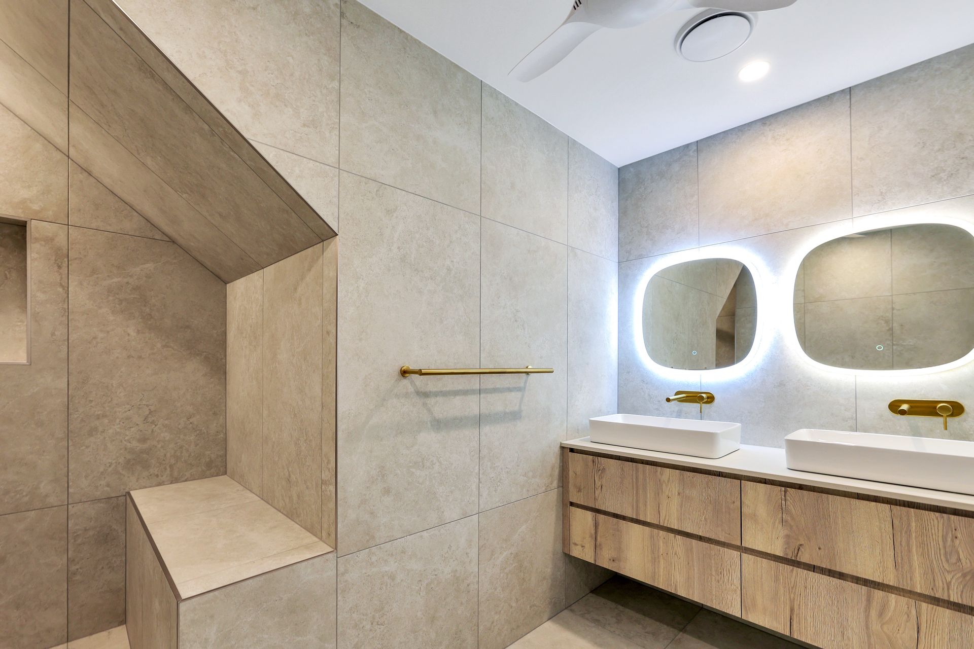 Modern bathroom with light wood vanity, dual sinks, and gray tile walls. Two mirrors with halo lighting.
