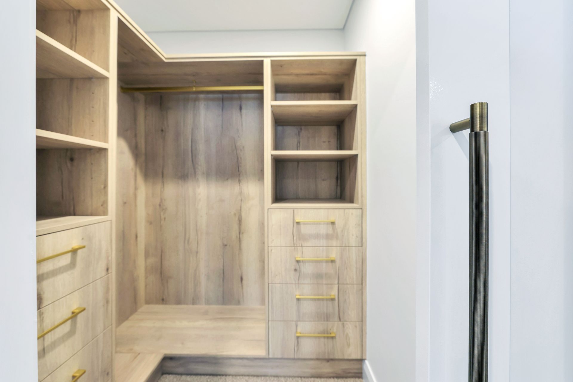 Walk-in closet with light wood shelves, drawers, and a hanging rod. Gold hardware. Pale walls.