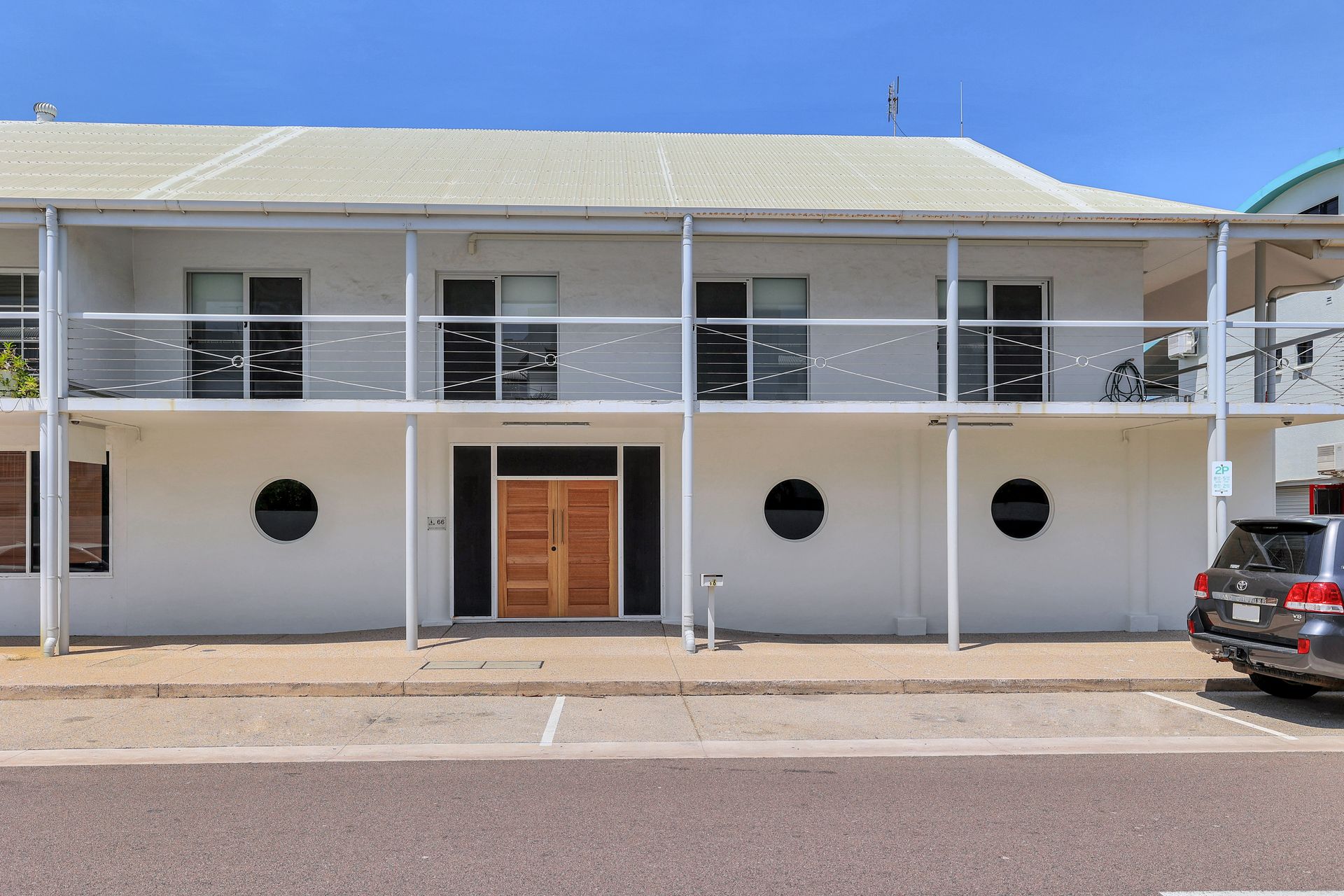 White two-story building with balcony, porthole windows, and wooden double doors. A car is parked in front.