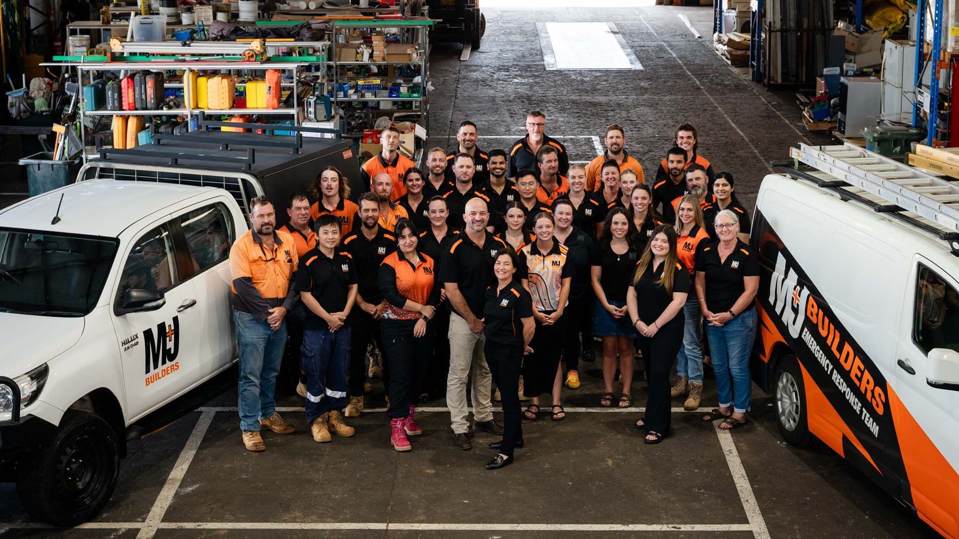 More than 30 M+J Builders team members in a large warehouse between two M+J Builders branded vehicles.