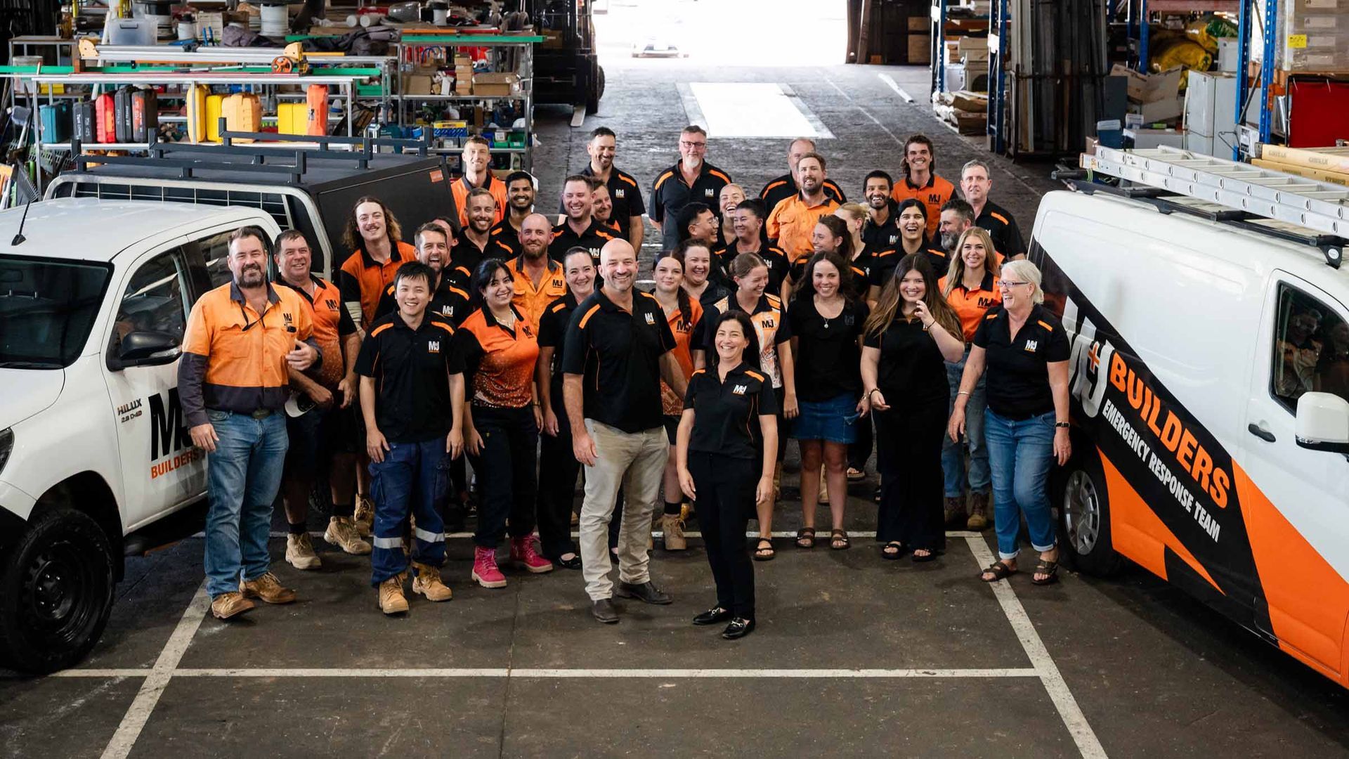 Group of builders in orange/black shirts, standing near work trucks inside a warehouse.