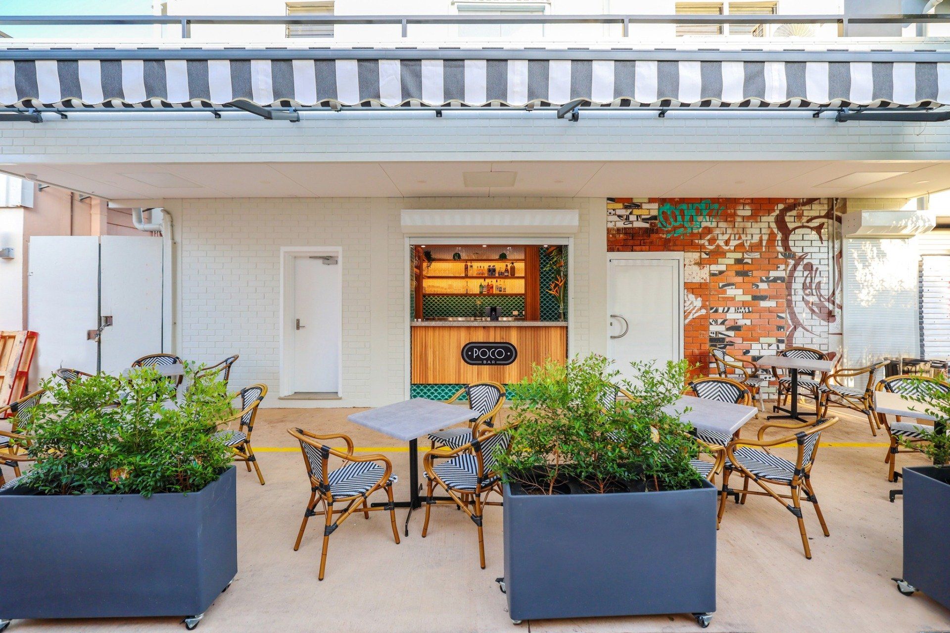 Outdoor cafe with tables, chairs, and potted plants, under a striped awning and mural-covered wall.