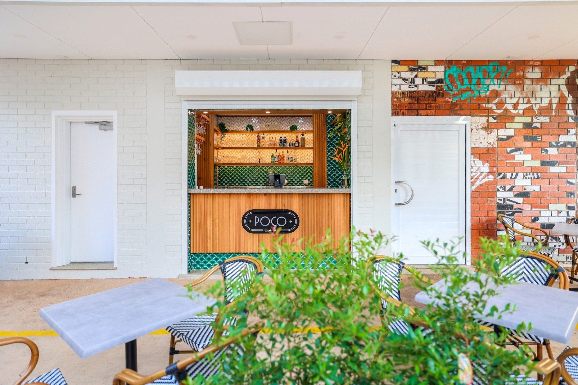 Outdoor bar with wooden counter, green tiles, and roll-up shutter. Tables with checkered chairs are in front.