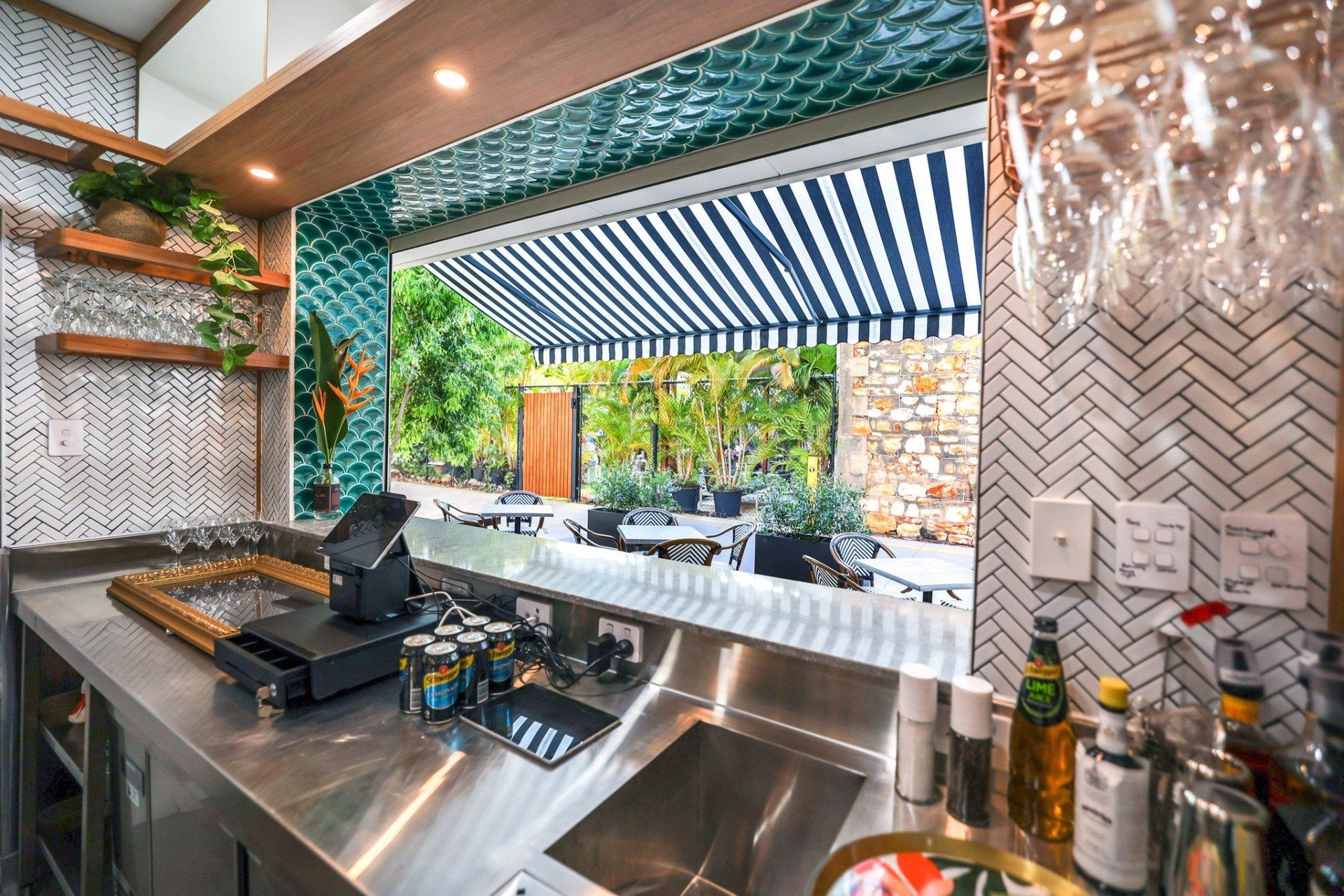 Bar interior with stainless steel countertop, open to outdoor seating under a striped awning.