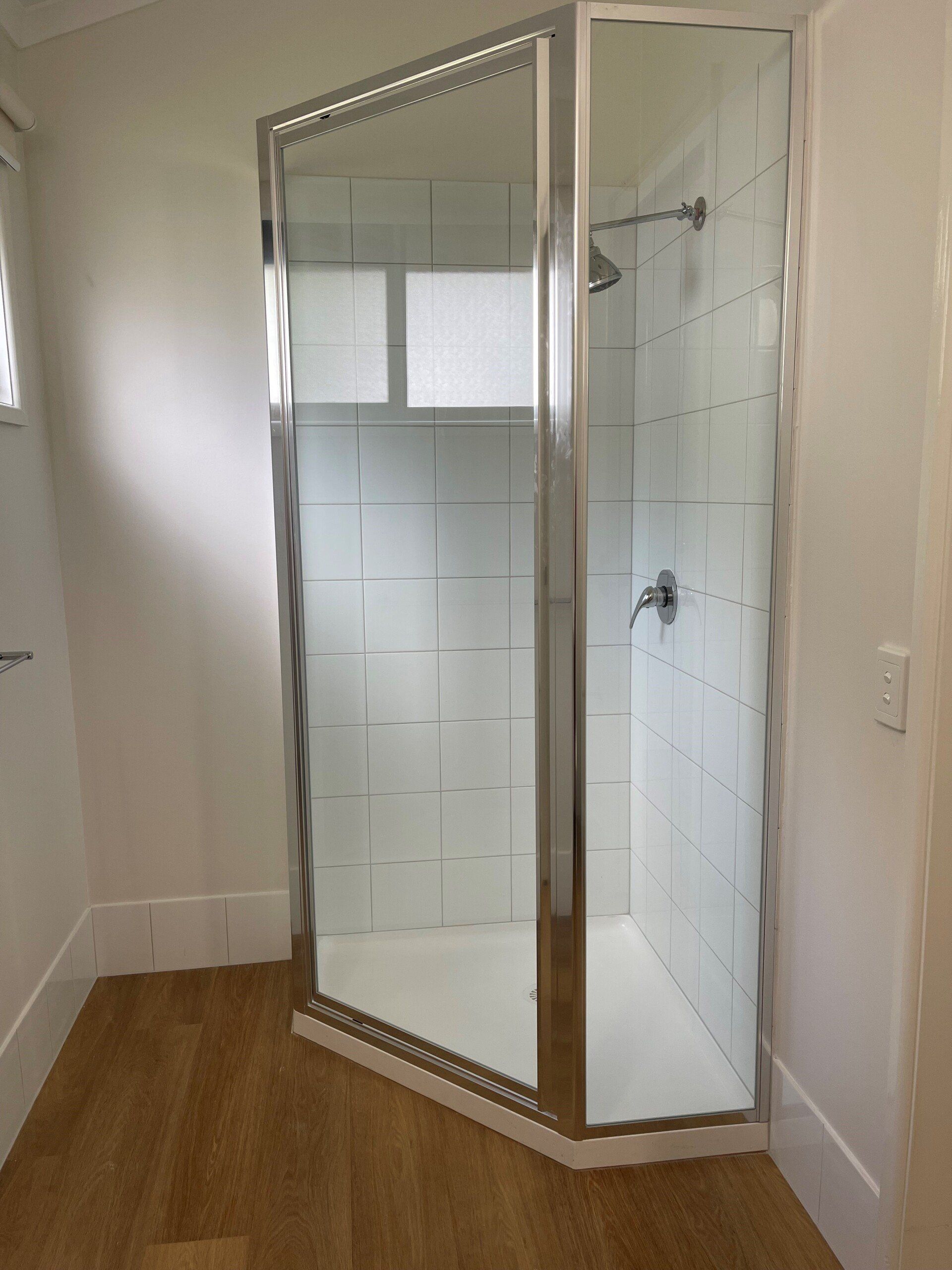 A corner shower with glass doors, chrome frame, and white tile interior. Light wood floor and white walls.