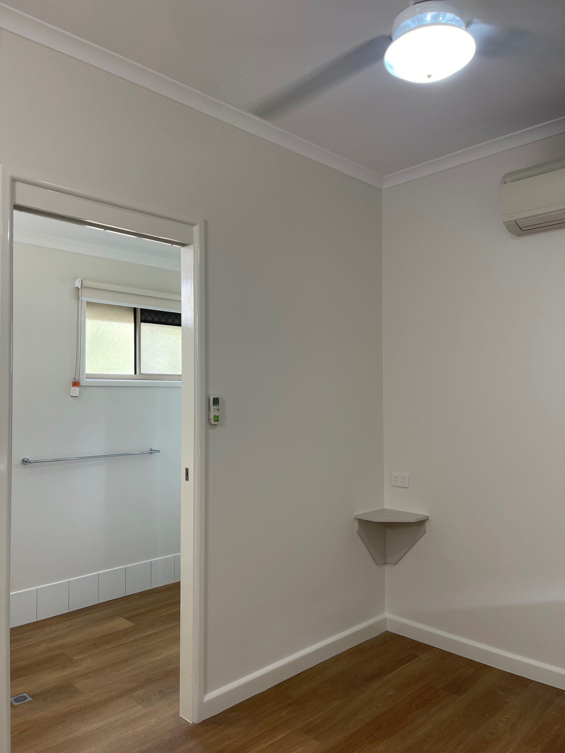 Empty white room with open doorway to a bathroom. Wood floors, ceiling fan, and a small shelf in the corner.