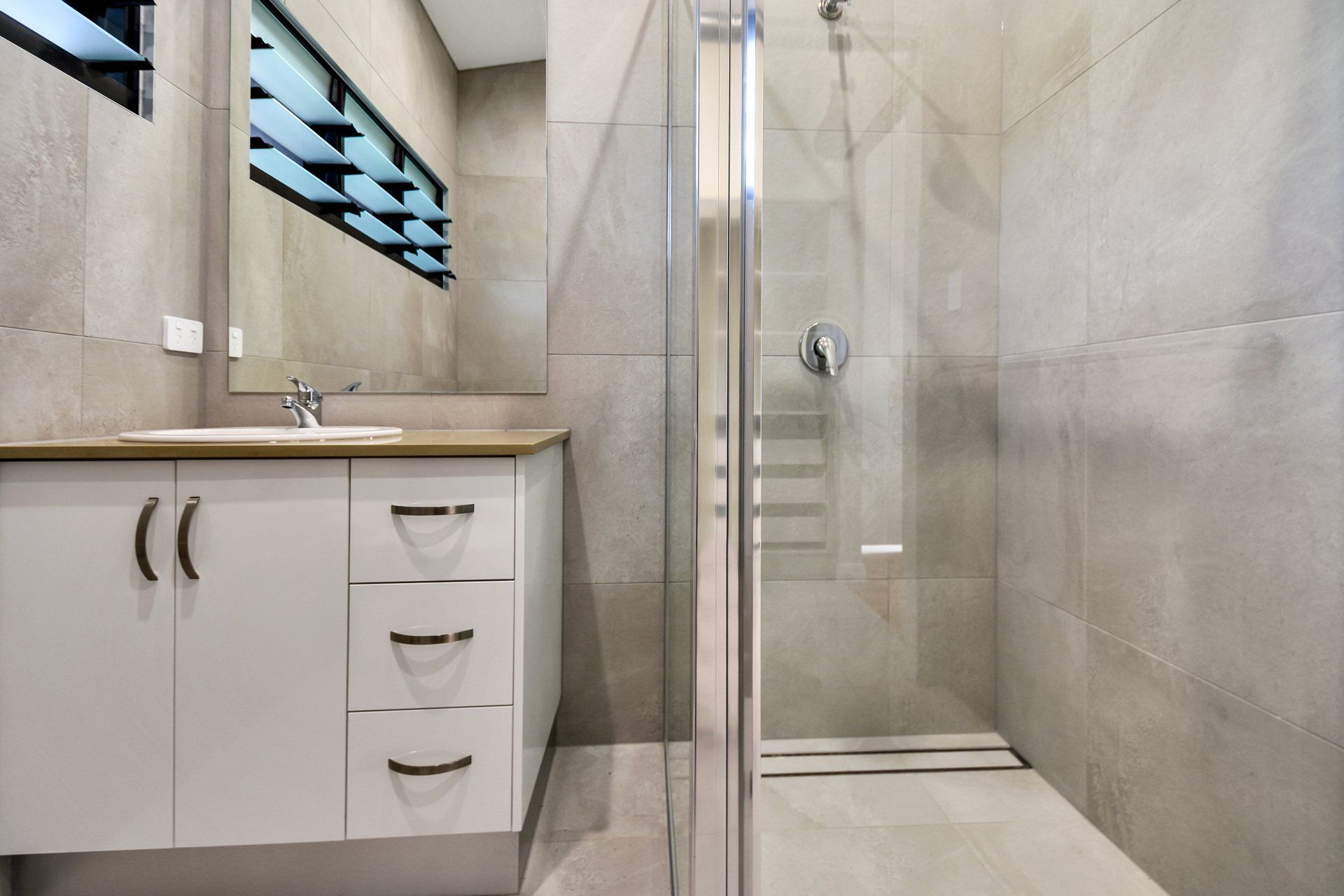 Bathroom with white vanity, mirror, and glass shower. Light gray tiled walls and floor.