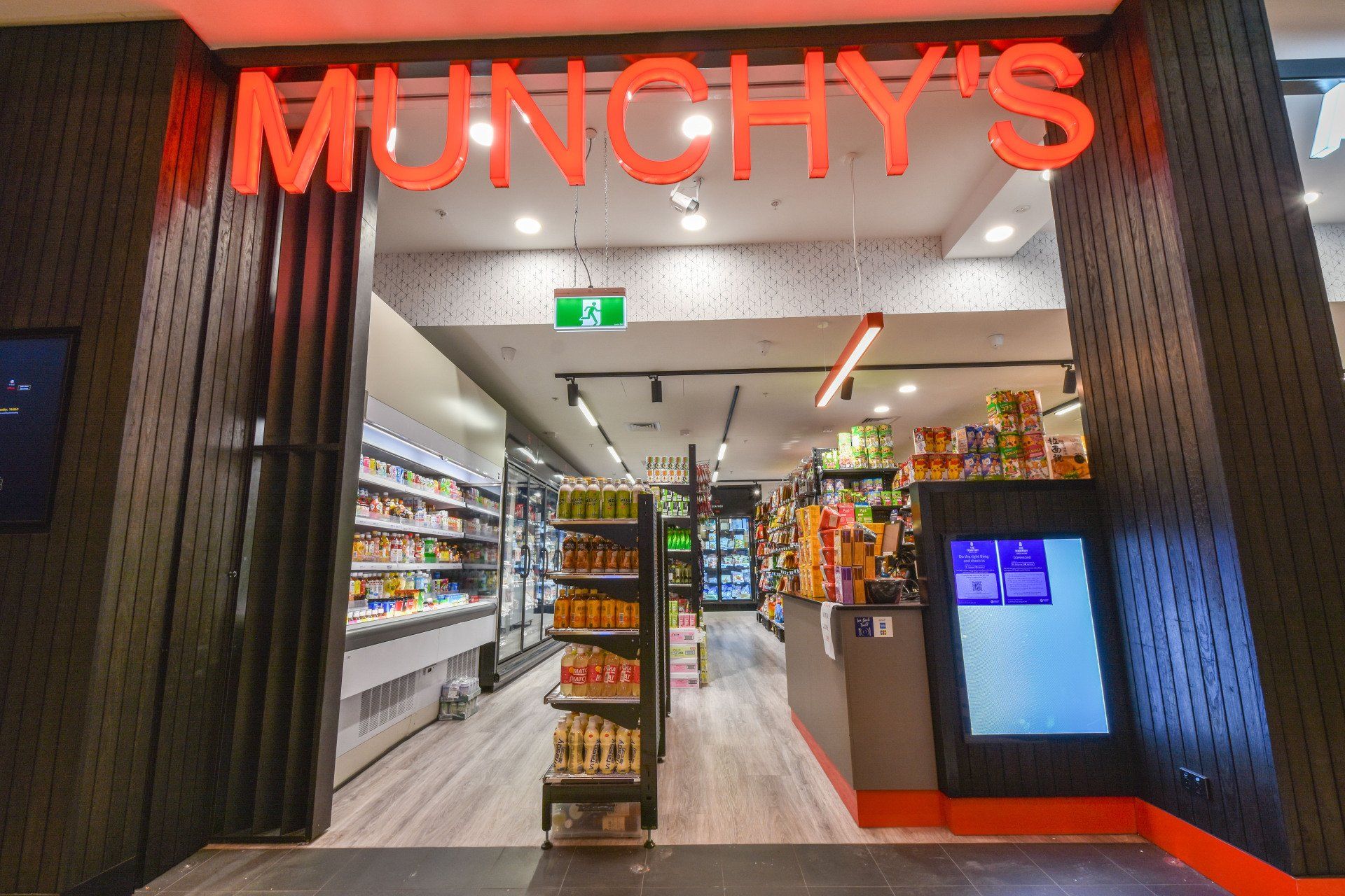 Munchy's store entrance with red neon sign. Inside, shelves of snacks and drinks are visible.