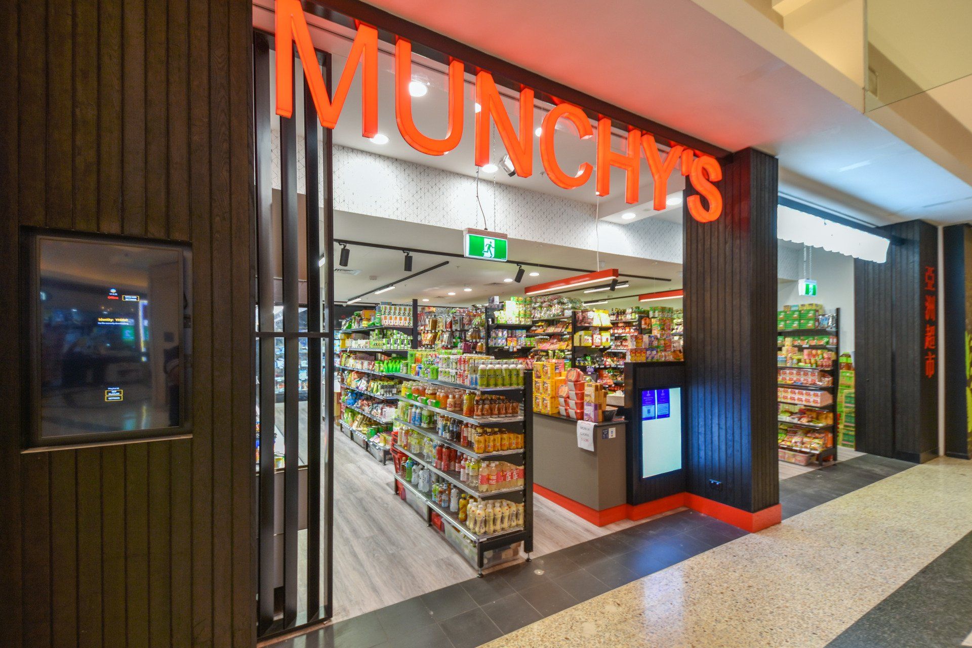 Munchy's convenience store entrance. Orange sign over doorway. Interior with shelves stocked with goods.