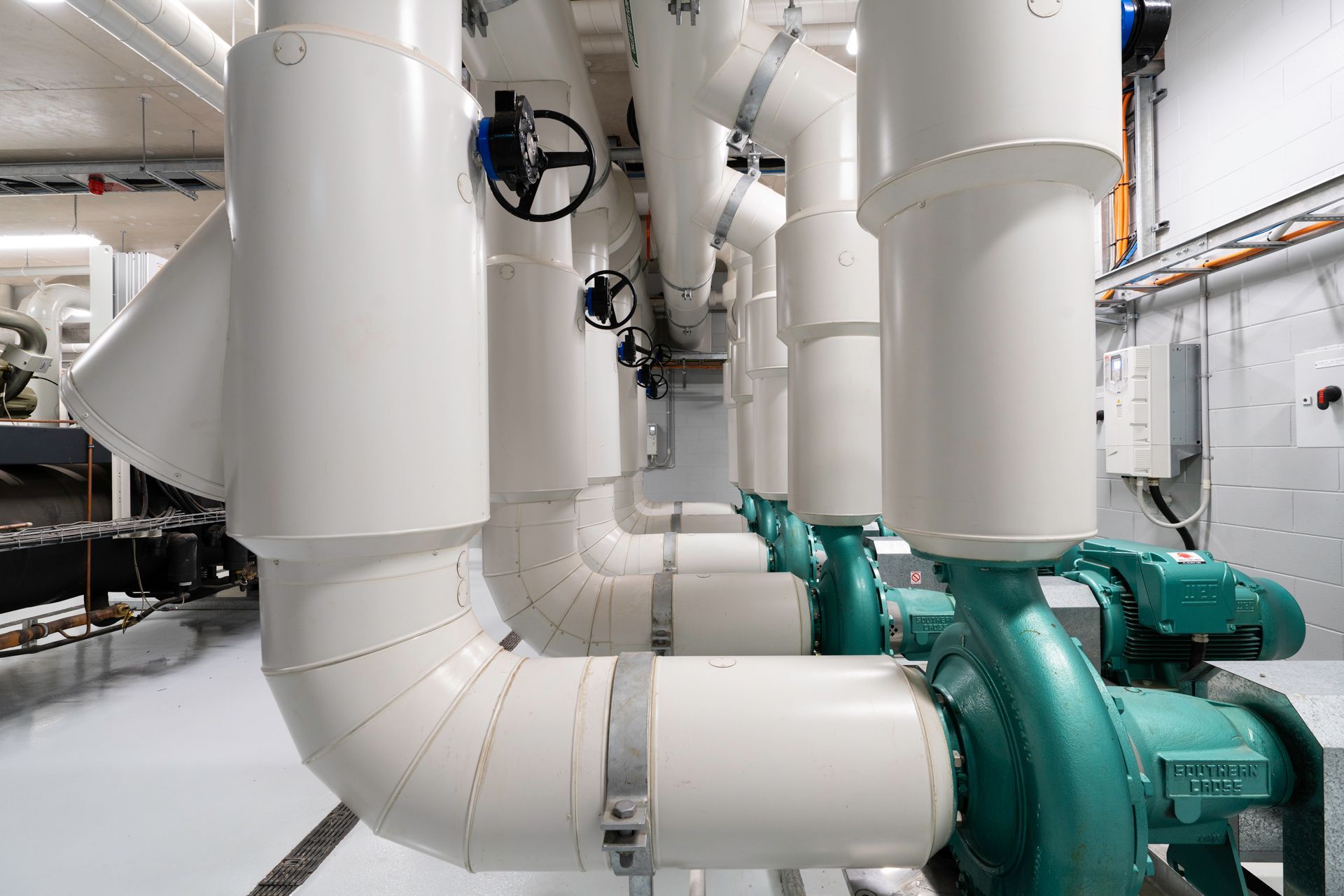Pipes and pumps in a mechanical room. White pipes, teal pumps, valves. Industrial setting.