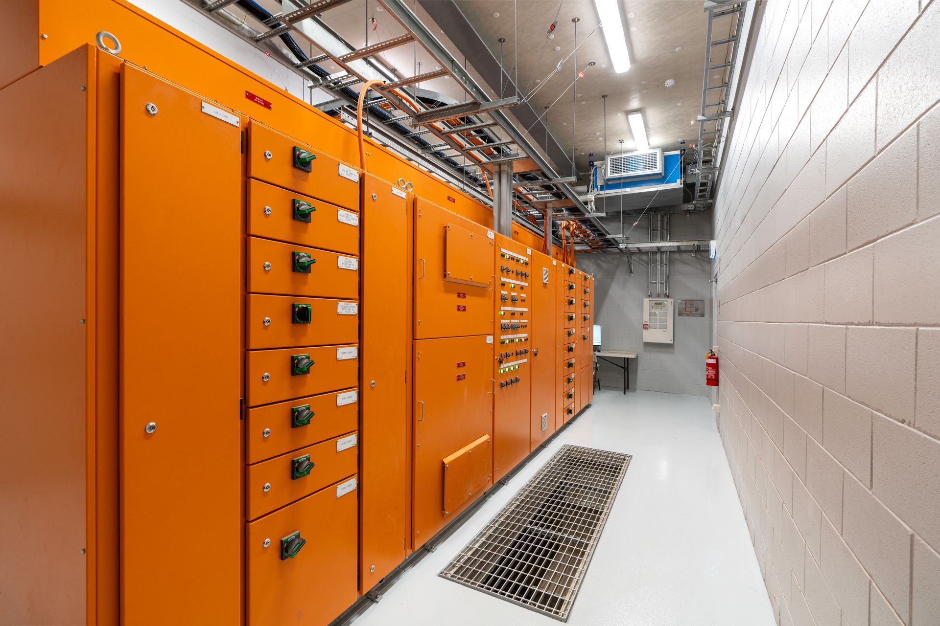 Orange electrical panels line a narrow room with white walls, copper pipes, and a floor grate.