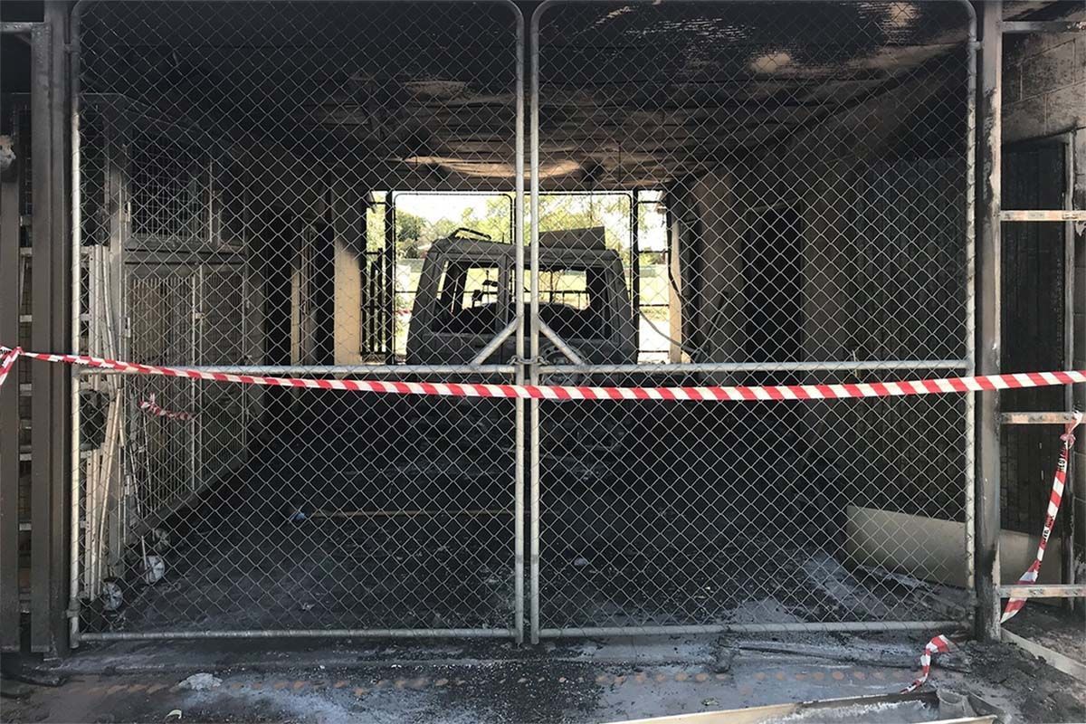 Burnt out vehicle behind chain link gate, red/white tape. Structure walls blackened with fire damage.