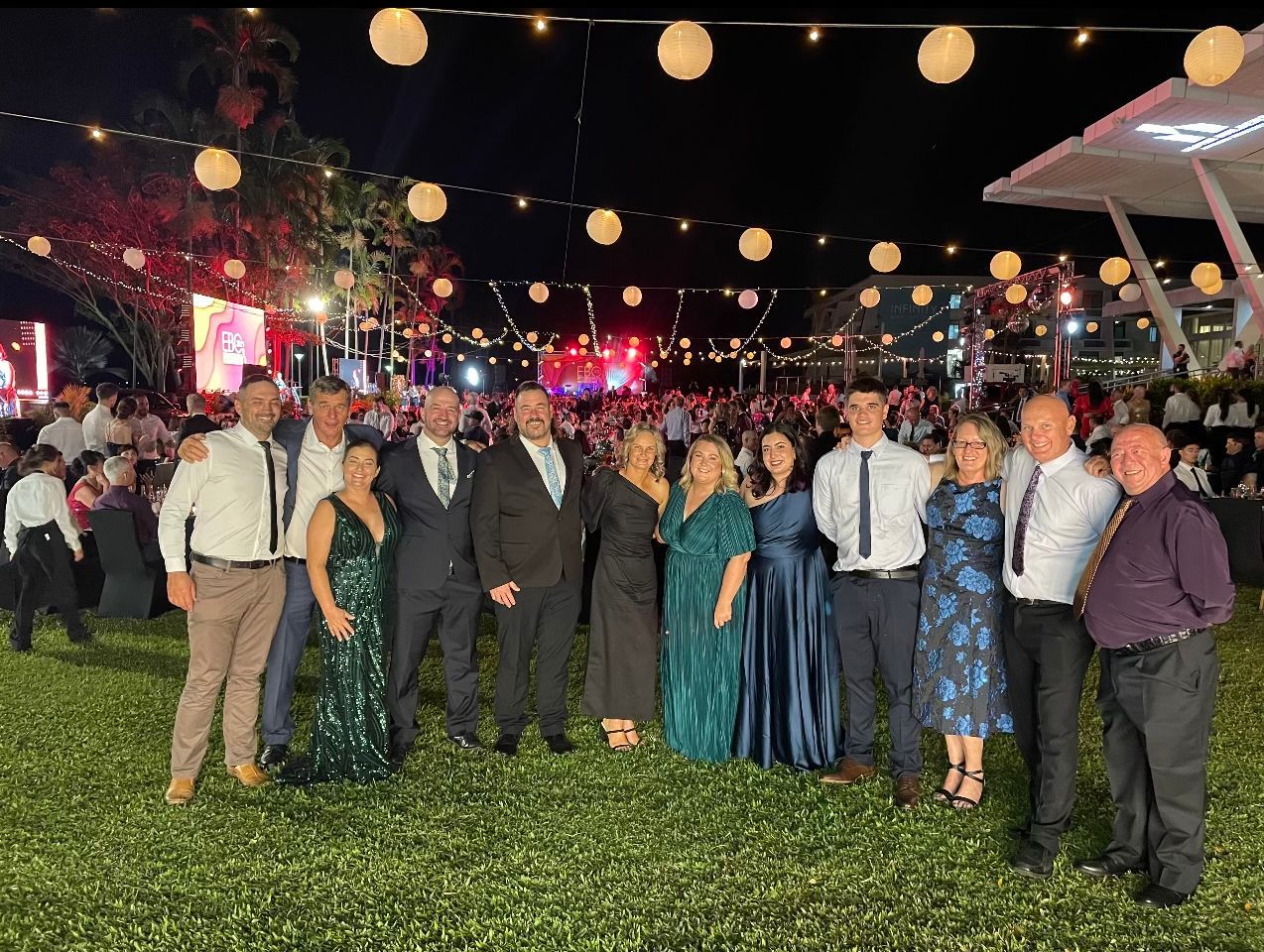 Group of people in formal attire pose outside at night, with string lights overhead.