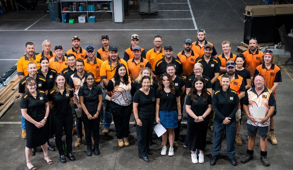 Group photo of a diverse team in a warehouse. Some in black shirts, others in orange. All are smiling.