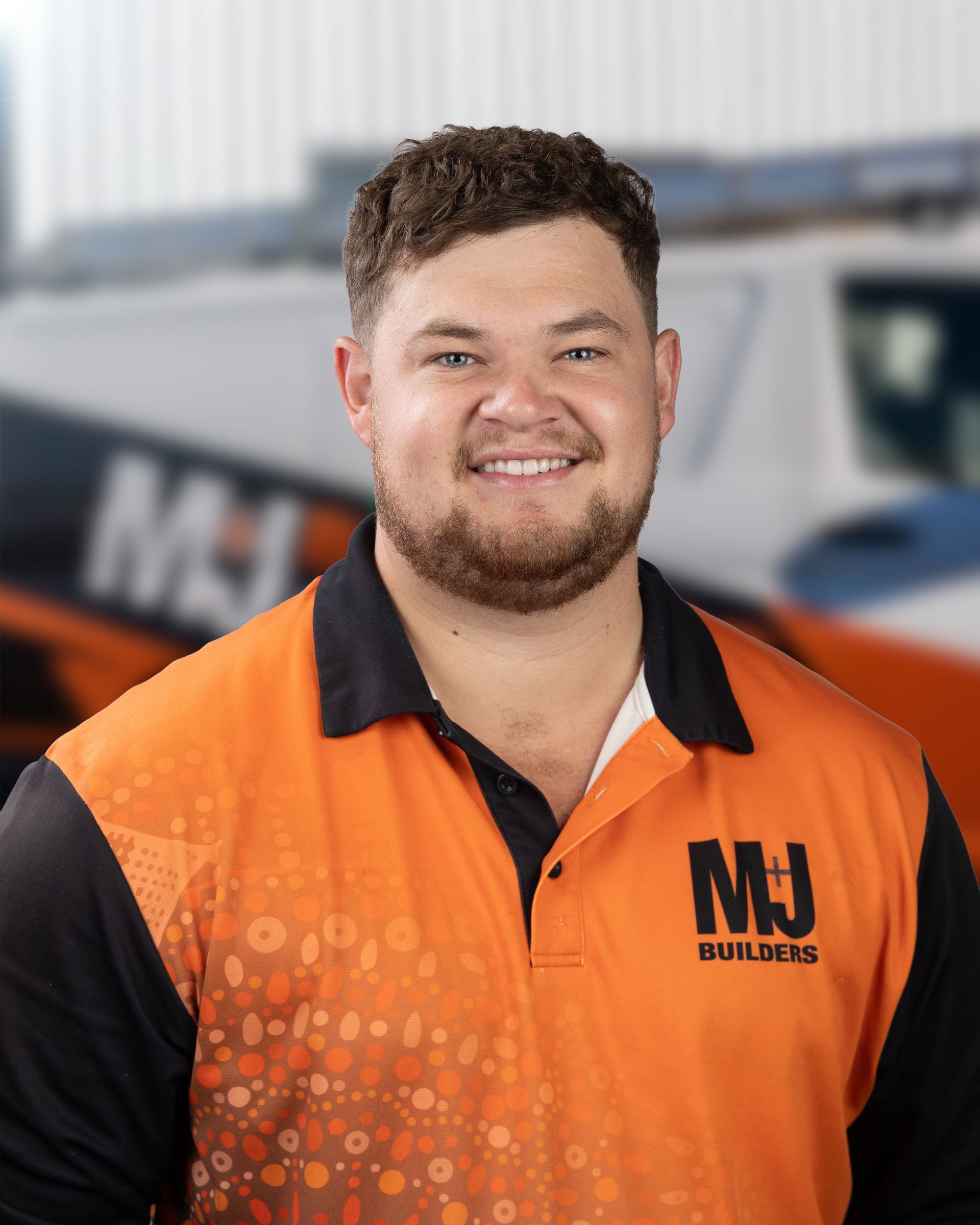 Man in orange work shirt smiles; blurred background of a truck and building.