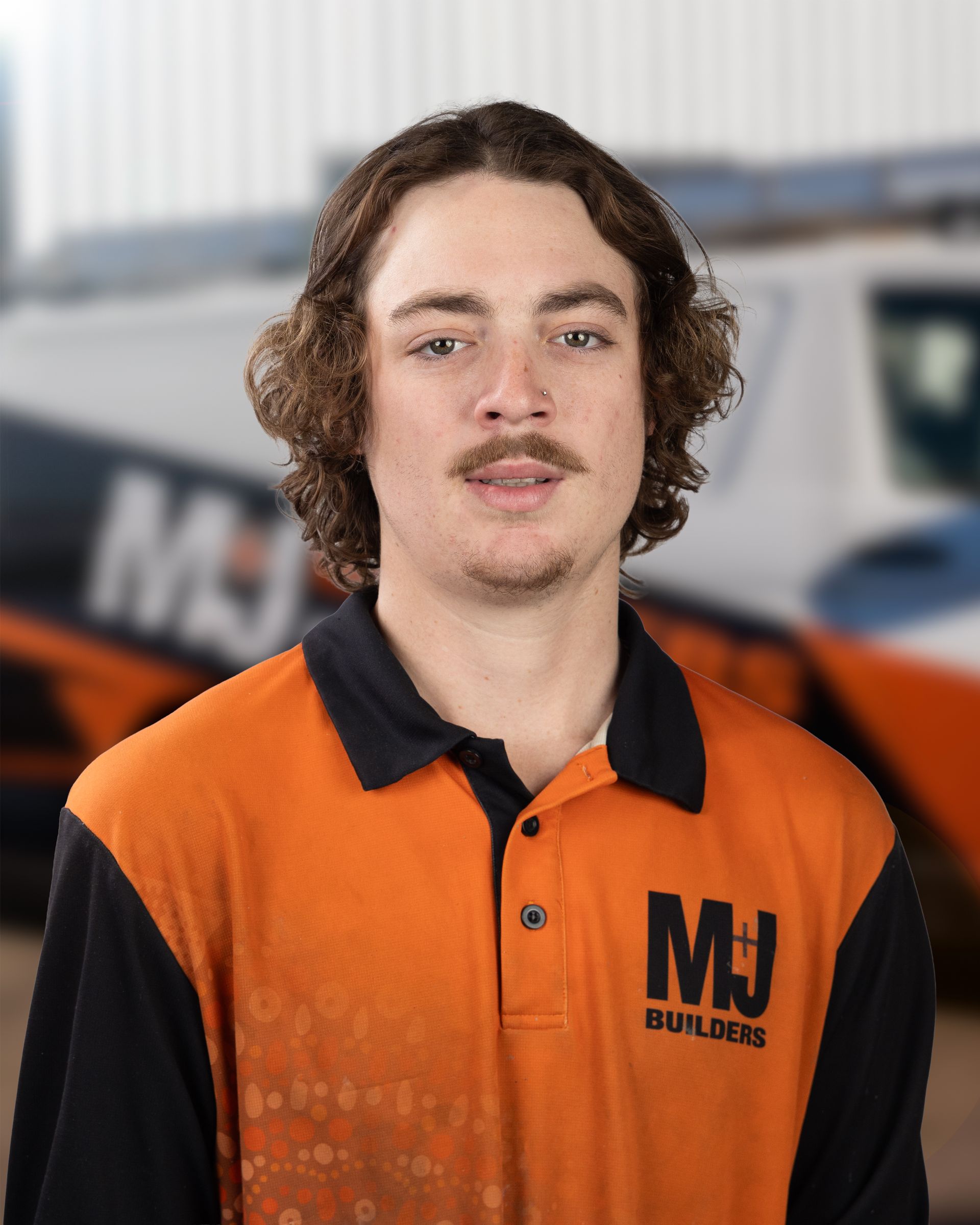 Young man with long curly hair in orange work shirt smiles on a construction site.