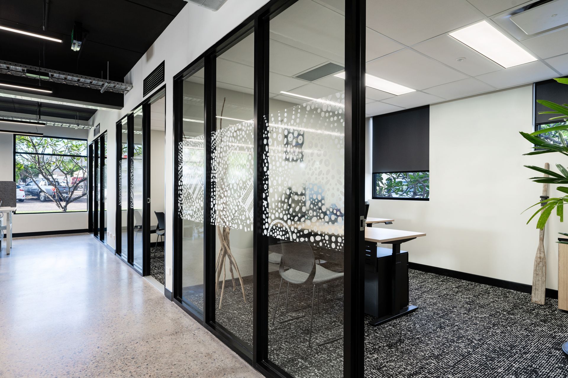 Office hallway with glass-walled rooms; black frames, speckled flooring, and black roller blinds visible.
