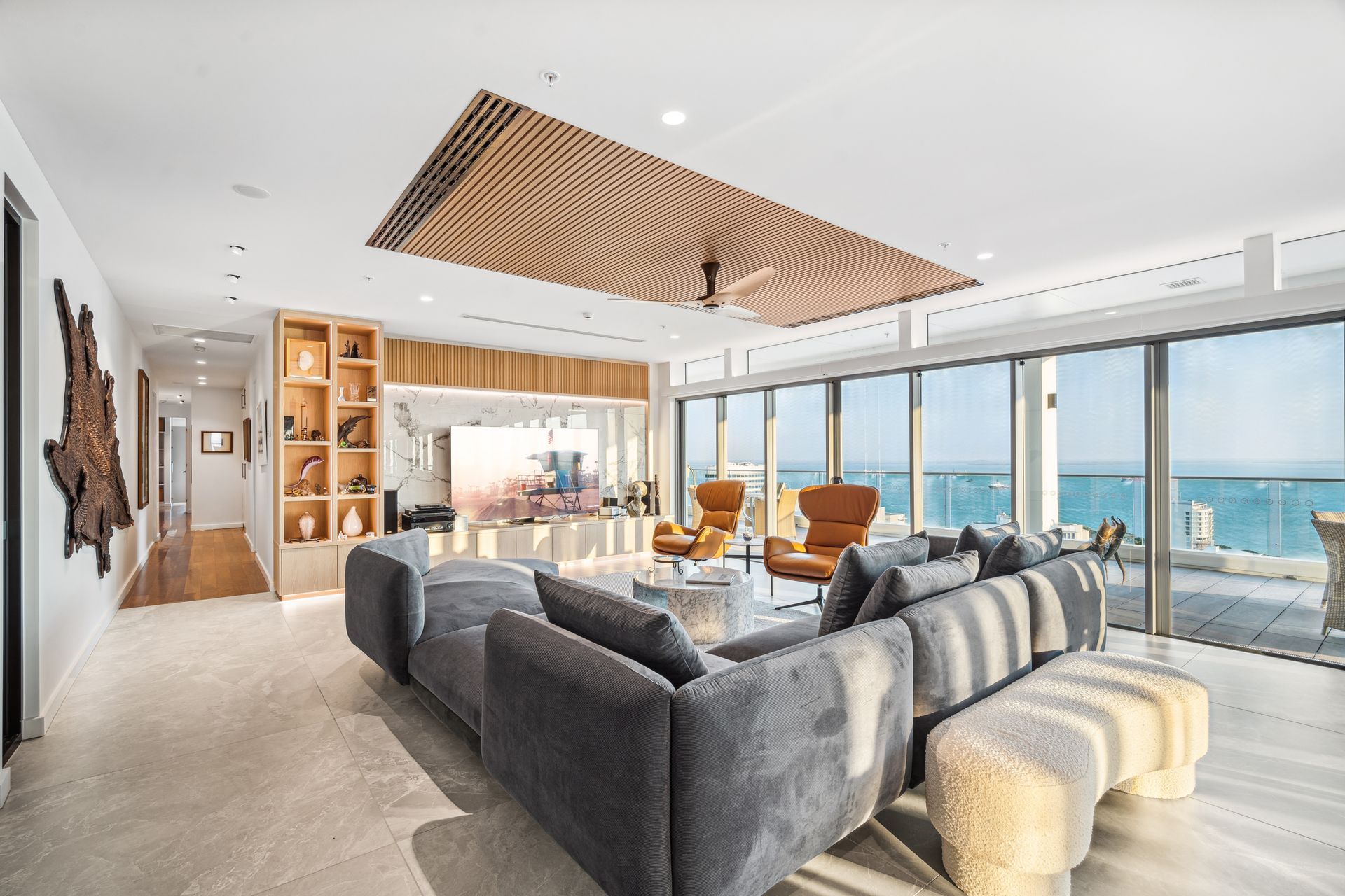 Residential living room with large marble floor tiles, custom wooden TV unit tv unit, marble backsplash and wall to ceiling windows looking out at balcony and ocean view. There is a large grey velvet couch and two orange 60's style chairs with a round marble coffee table.