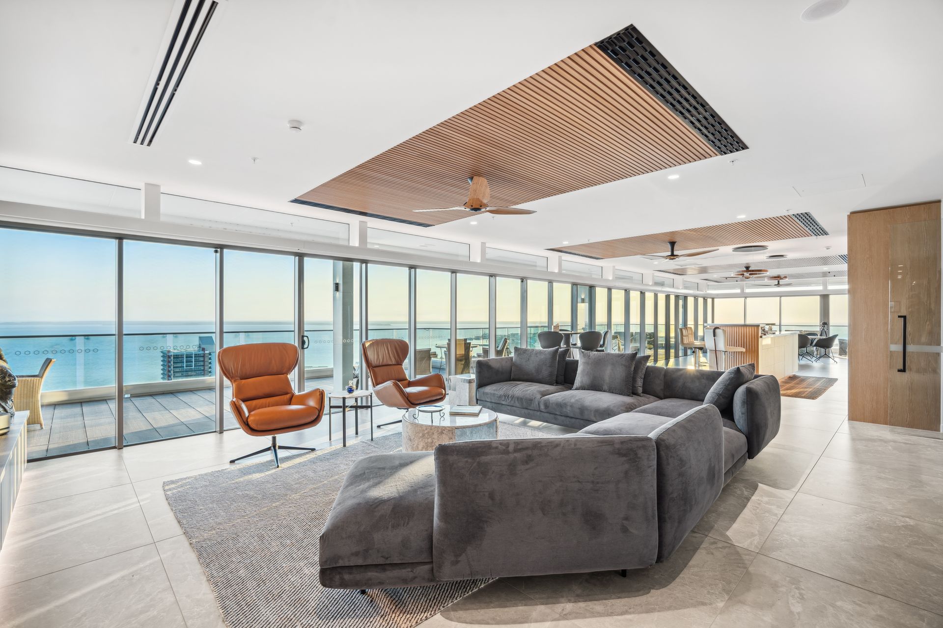 Spacious modern living room with ocean views, gray sectional sofa, leather chairs, wooden ceiling accents, and large windows.