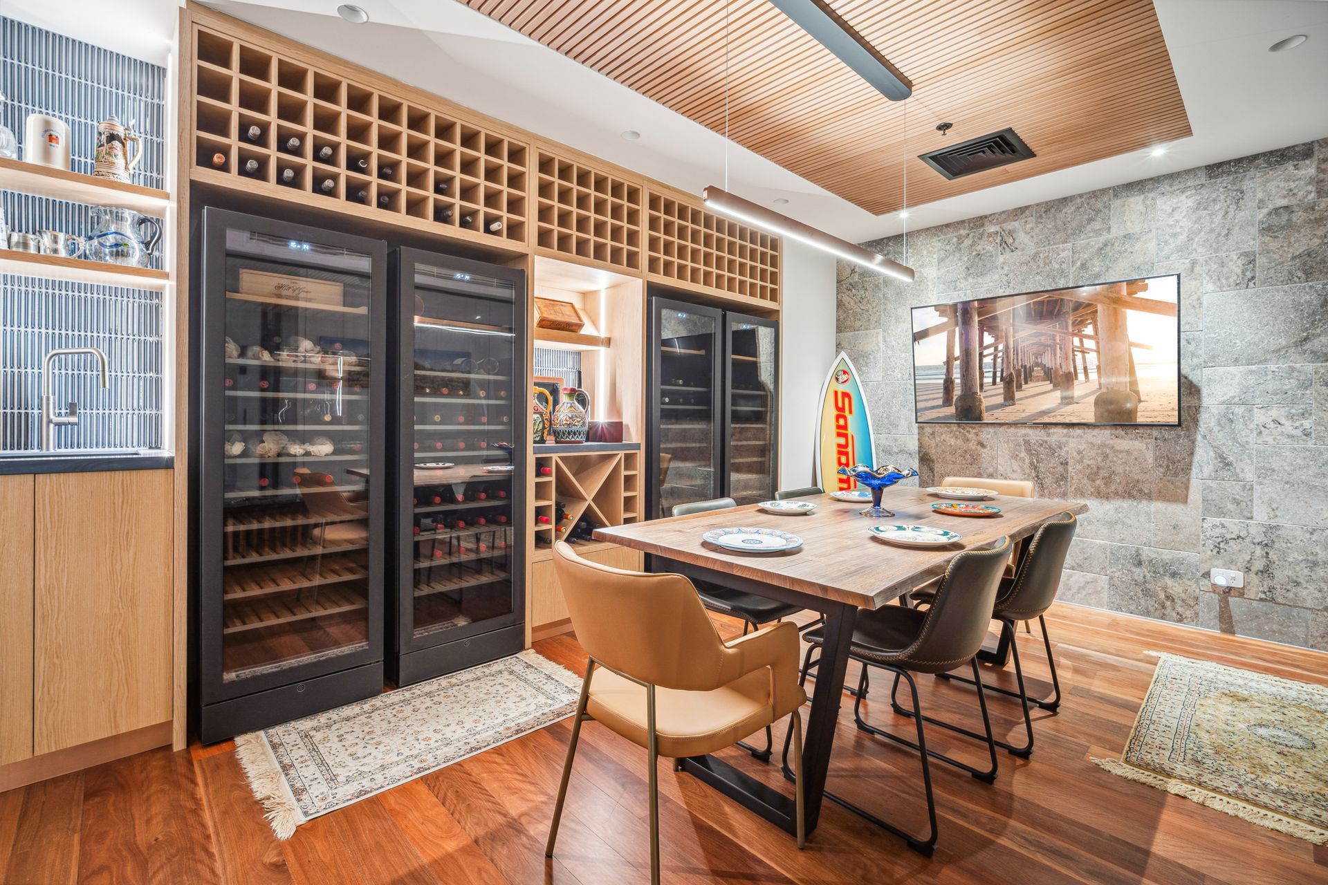 Residential dining area with wine racks and wine fridge, large stone tile wall, wooden floorboards and a colourful surfboard propped up in the corner.