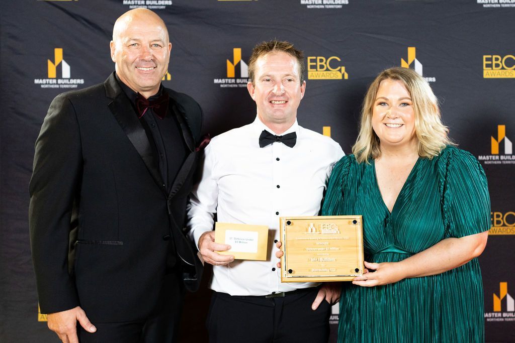 Three people holding awards, smiling at camera; black-tie event setting, gold and black backdrop.