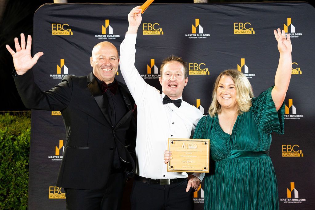 Three people smiling and celebrating, holding a plaque and award at an event with a gold and black backdrop.