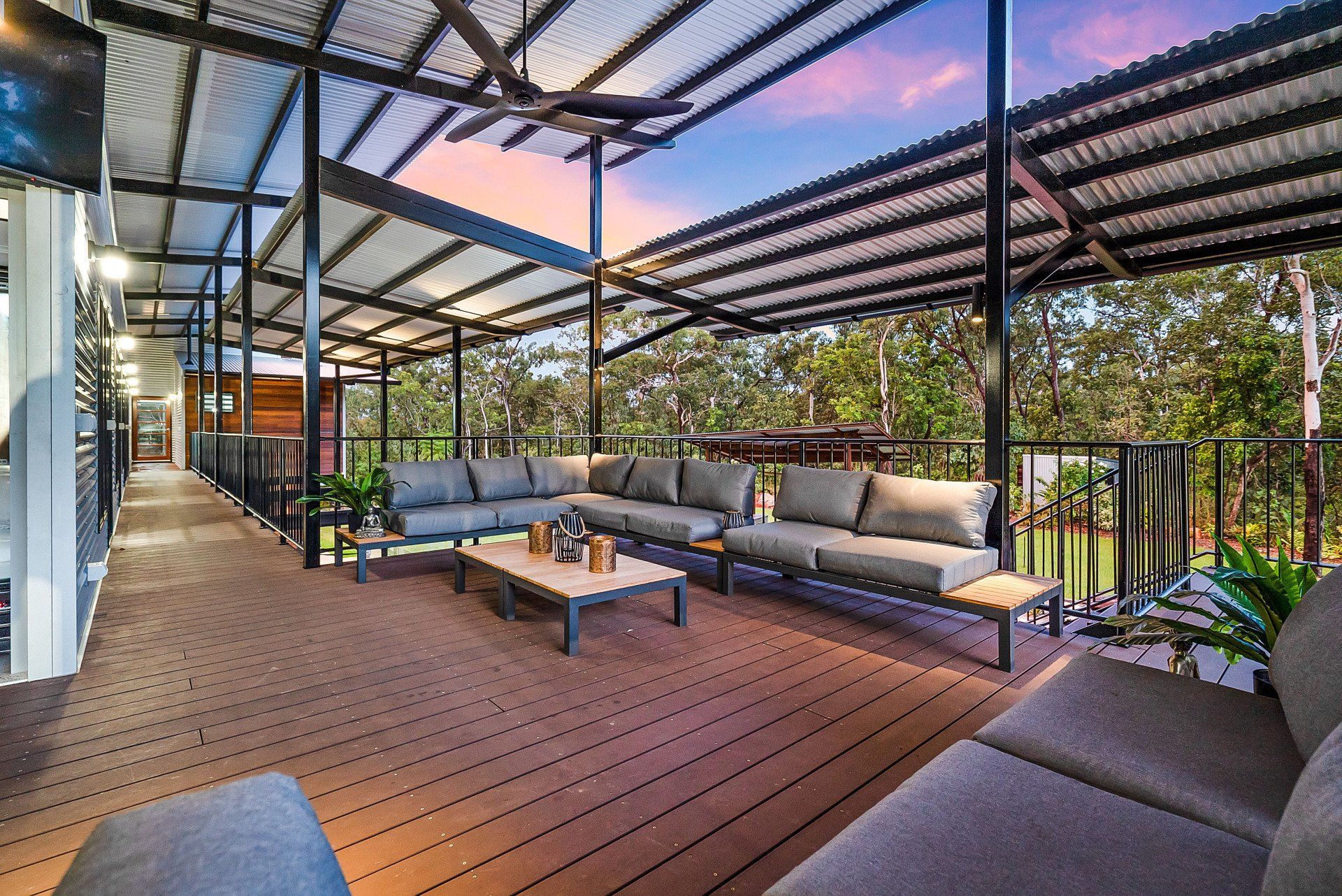 Outdoor deck with gray couches, table, and metal roof overlooking trees at dusk.