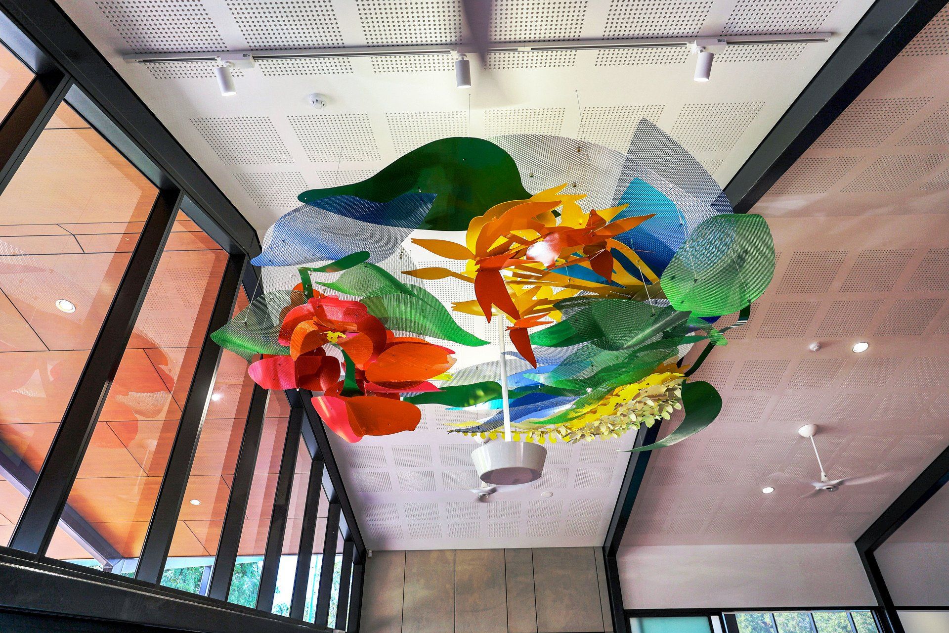 Colorful glass art installation in a building with high ceilings and large windows; vibrant hues of red, orange, and green.