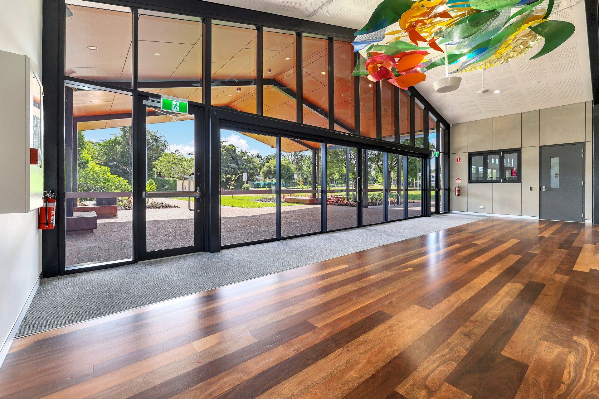 Hall with wooden floor, glass walls overlooking a park, and colorful artwork on ceiling.