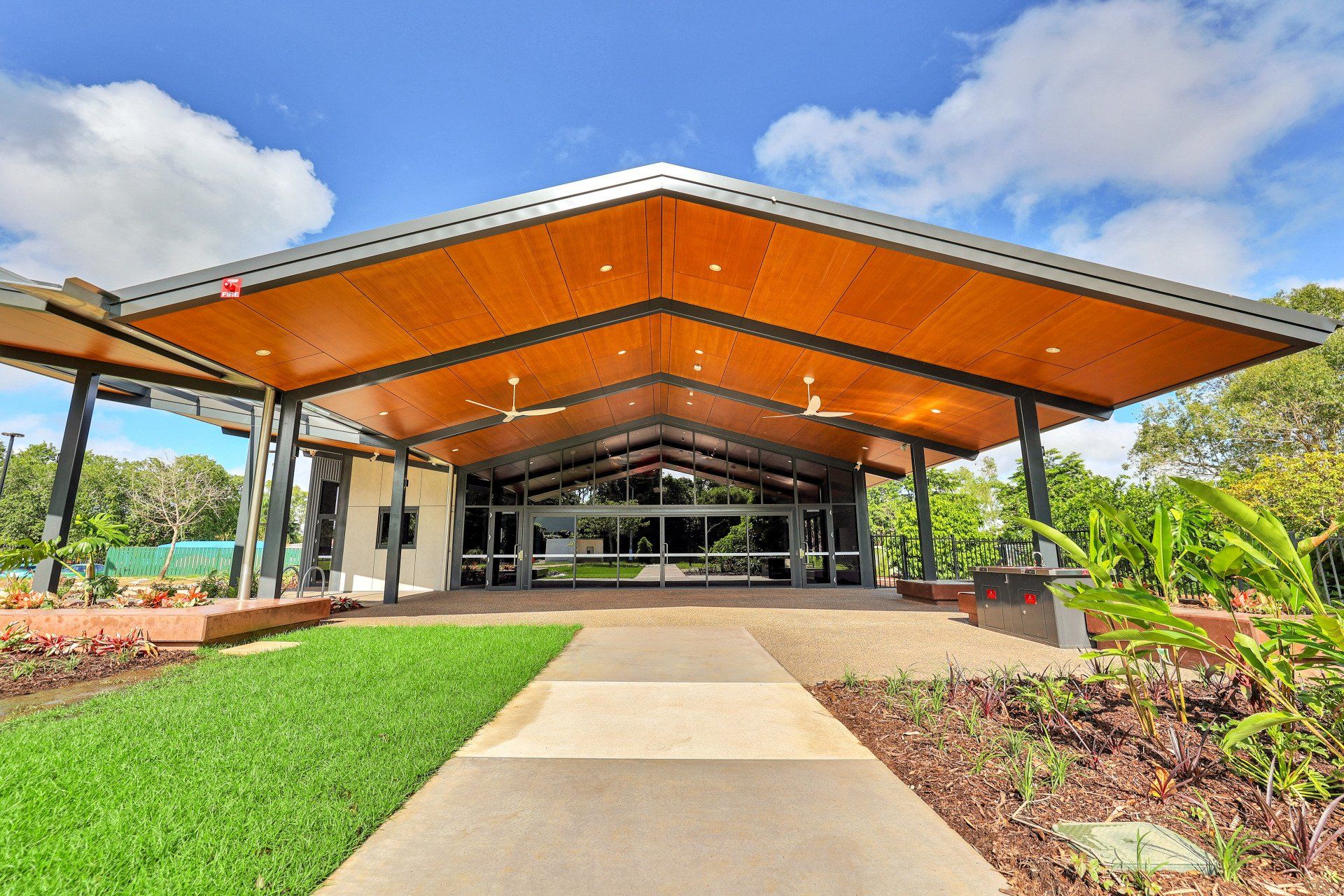 Large undercover area with barbeques, seating and garden leading to a community hall. Shaded by large roof with wooden panels, lights and fans.