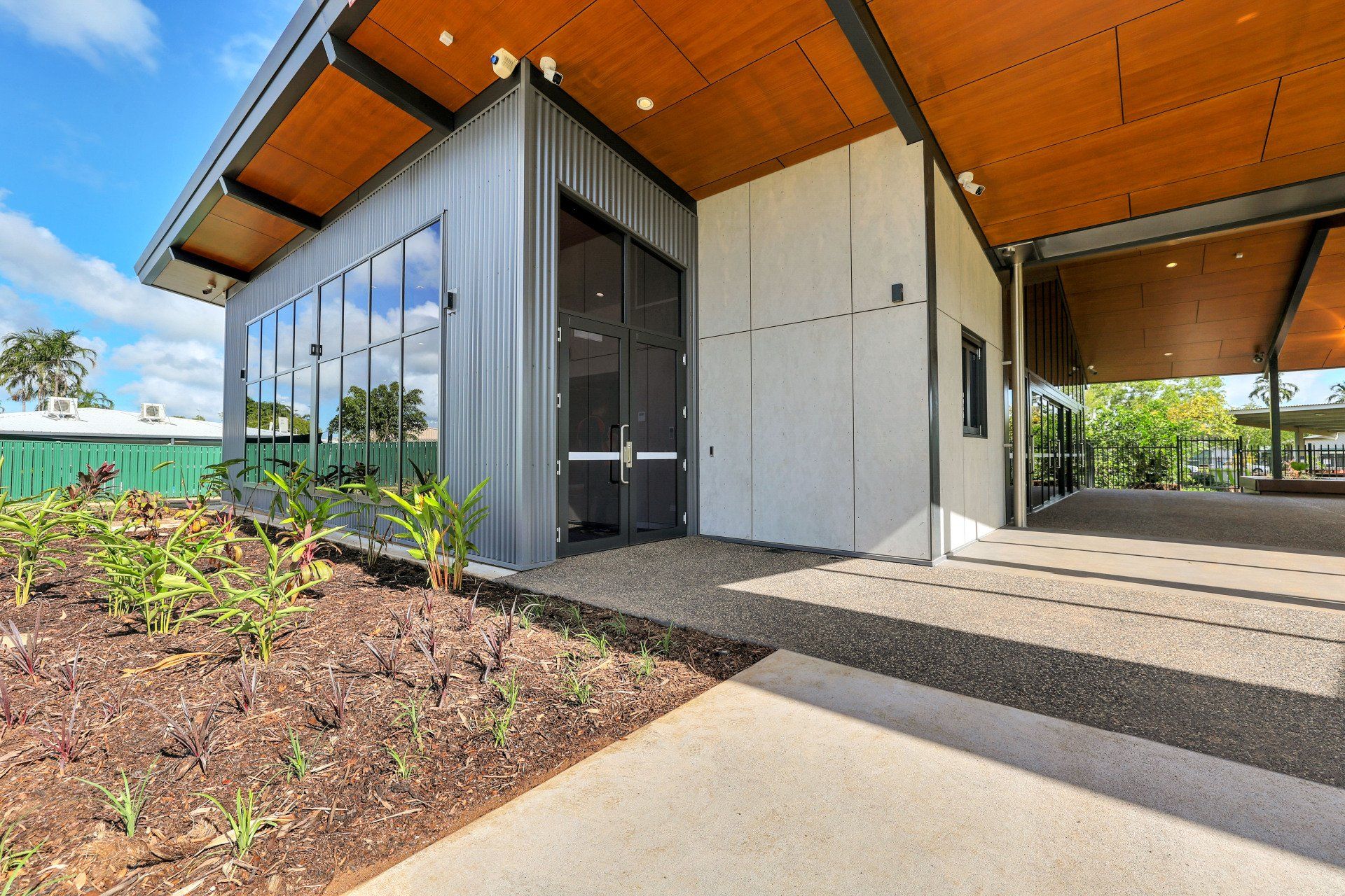 Modern building with a corrugated metal facade, large windows, and a covered walkway; blue sky overhead.