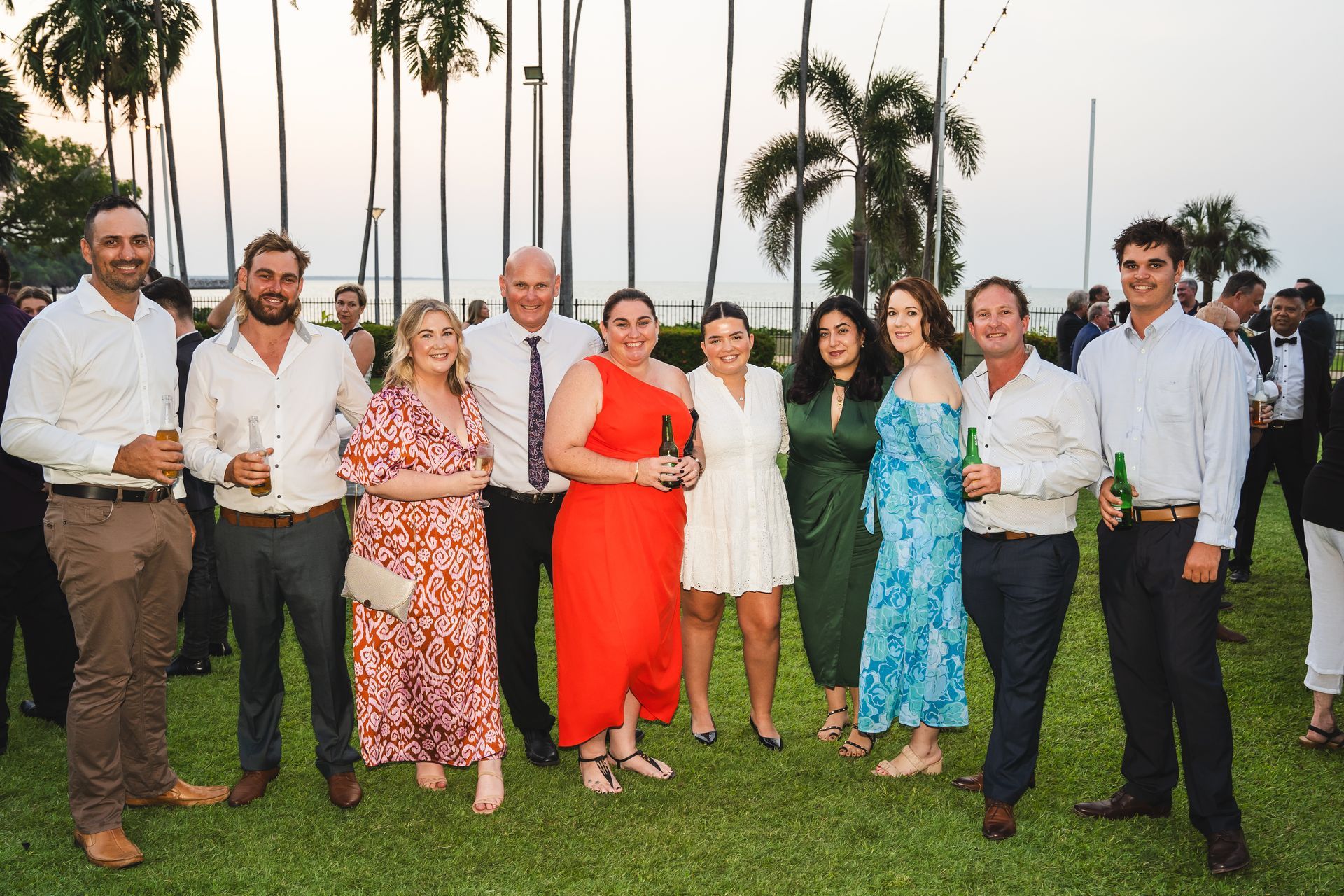 Group of people smiling, holding drinks at an outdoor event on grass with palm trees.