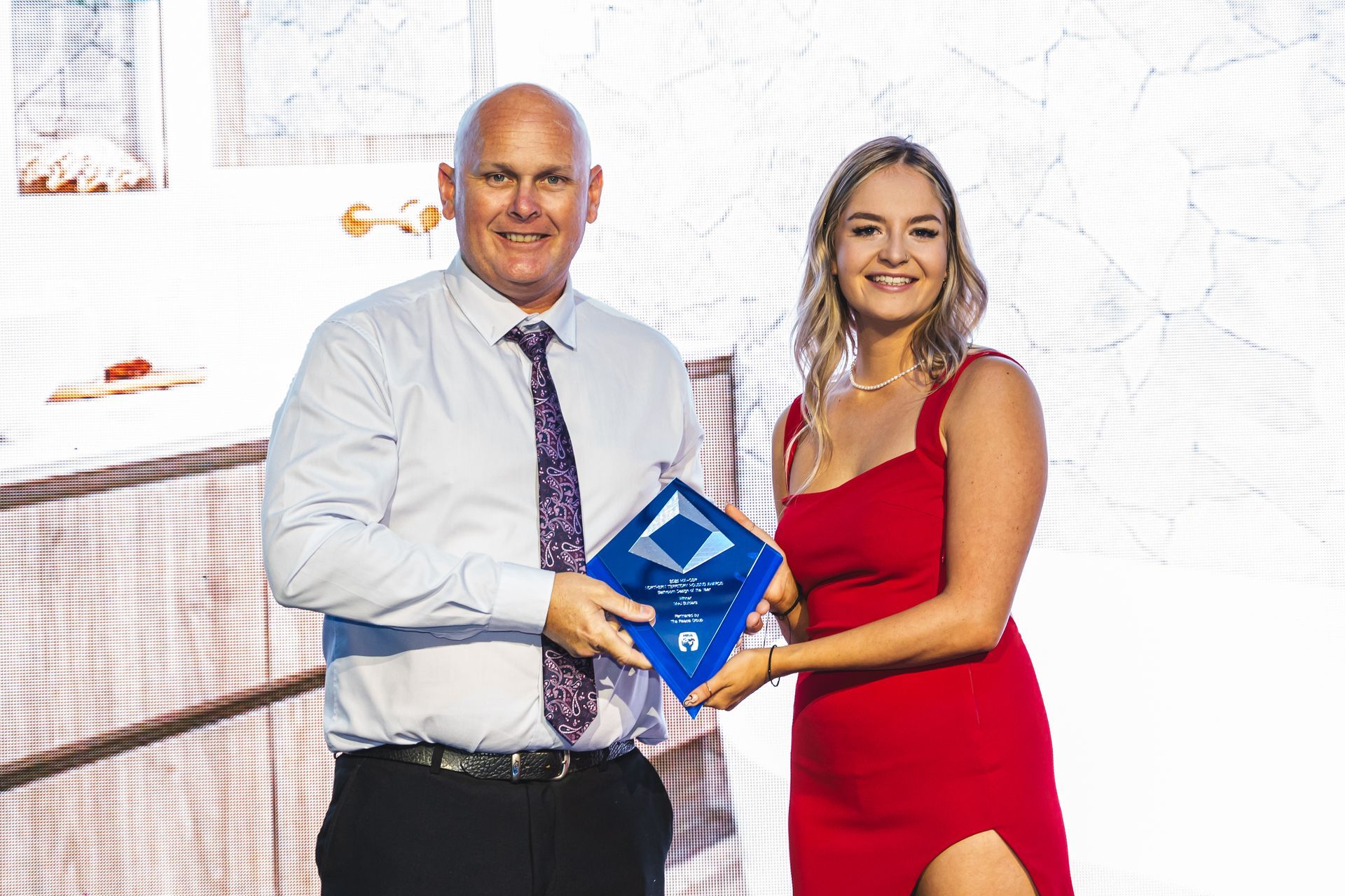Man in tie presenting an award to a woman in a red dress; outdoor event.