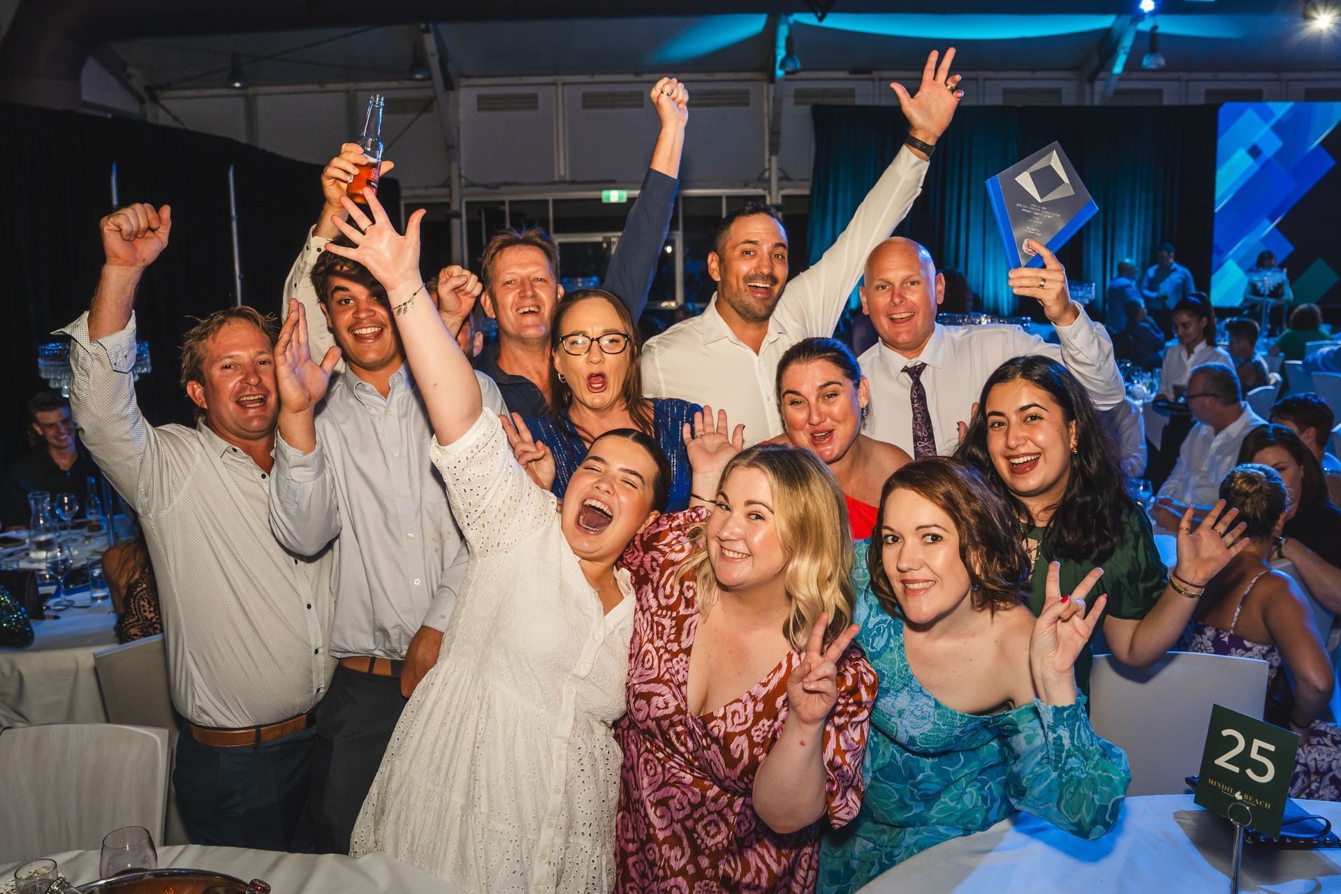 Group of people celebrating at an awards event, raising arms and smiling. Setting is a banquet hall.