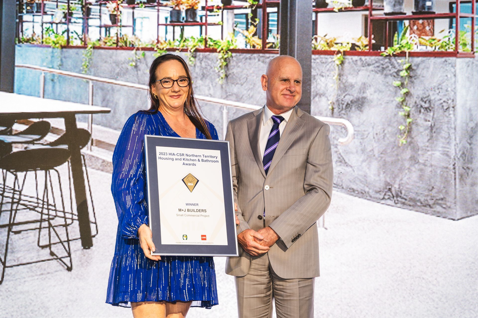 Woman in blue dress and man in suit holding framed award, interior setting with greenery.